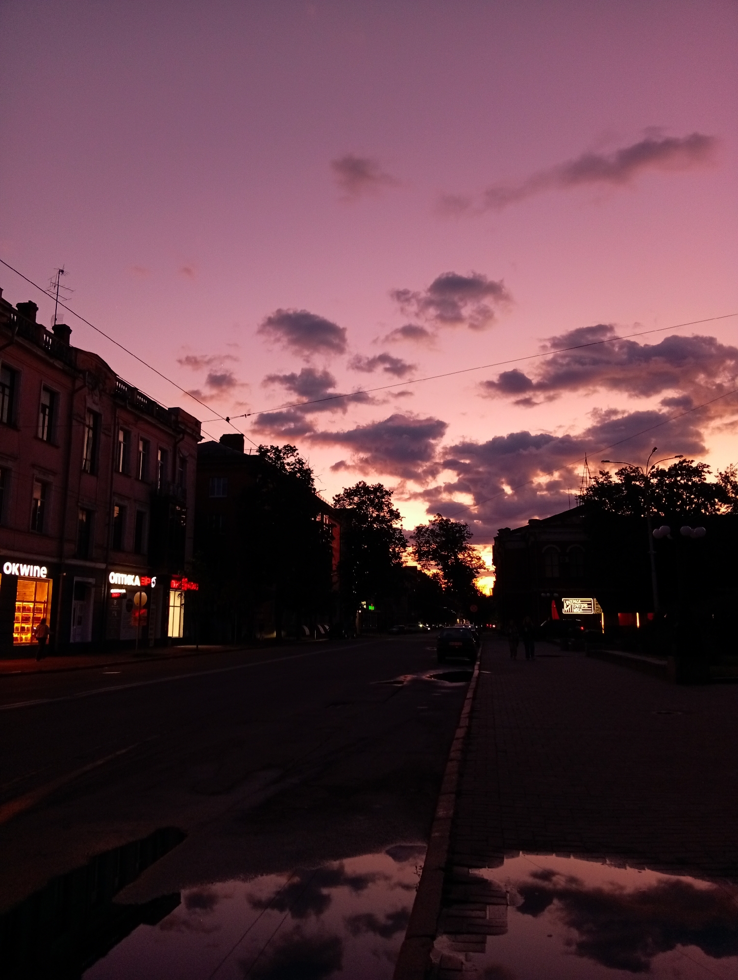 The image depicts an urban street scene in Poltava, Ukraine, at dusk or shortly after sunset, characterized by a striking pink and purple sky. Dark, scattered clouds are visible against the vibrant backdrop, some catching the last light of the day.
On the left side of the street, multi-story buildings with classical architecture line the road. Ground-floor shops are illuminated, with visible signs including "OKwine" (a wine store) and "ОПТИКА ЗОР" (Optics ZOR). A person can be seen near the entrance of the "OKwine" store, bathed in its warm light. An antenna is mounted on the rooftop of one of these buildings, and power lines stretch across the frame.
The street and sidewalk are wet, with several puddles prominently reflecting the colorful sky and the dark outlines of the buildings and clouds, adding a sense of depth and atmosphere. On the right side of the street, buildings and a line of trees are largely silhouetted, obscuring most details but revealing some distant illuminated signs. Two figures are visible walking on the right-hand sidewalk. Cars are parked along the street, and streetlights are beginning to cast their glow in the fading light. The overall scene evokes a peaceful, contemplative mood of a city transitioning into night.