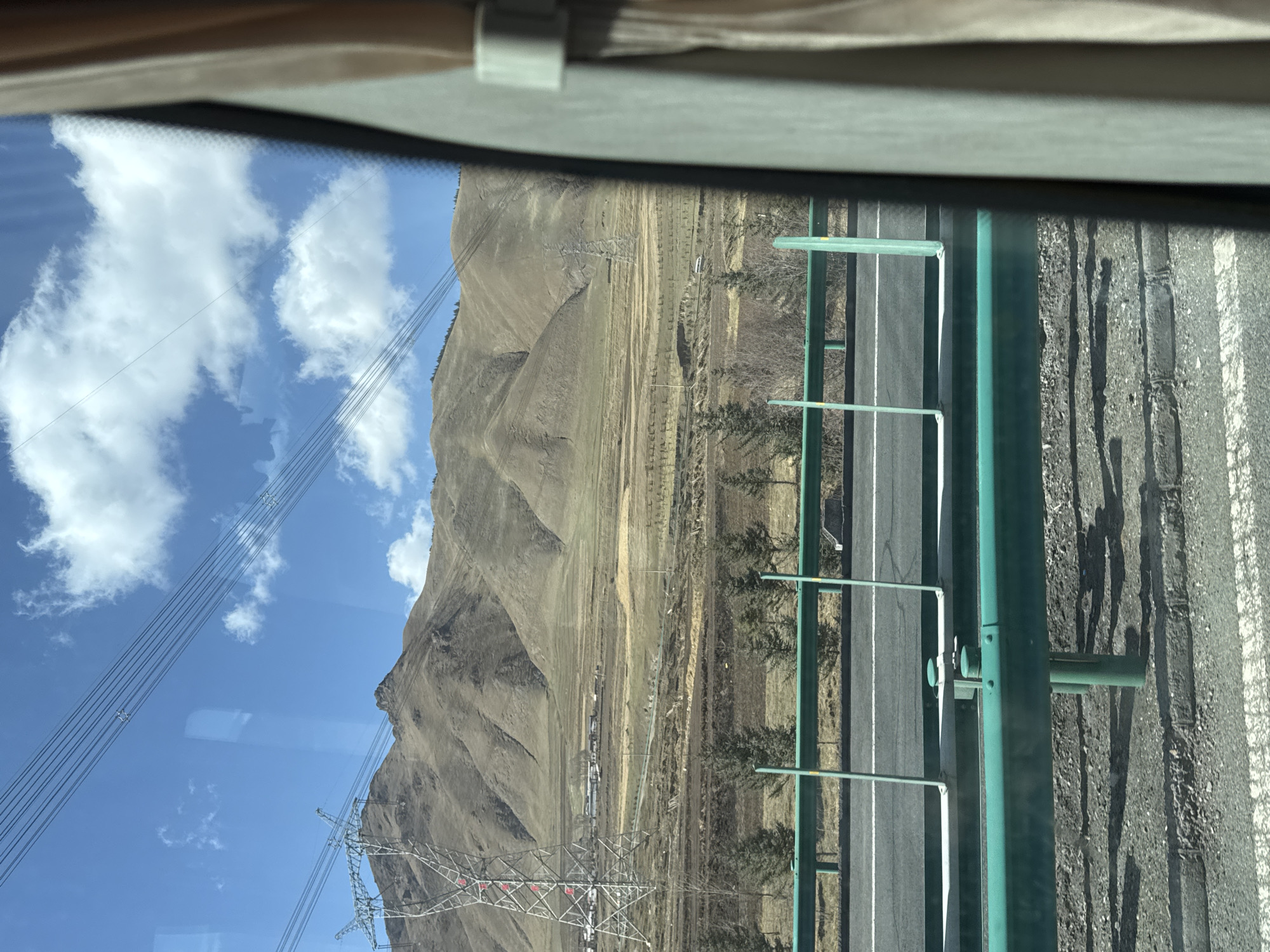 The image shows a view from inside a vehicle, likely a bus or car, looking out a window. The foreground contains a portion of the vehicle's interior, including a section of the window frame. Outside the window, a mountain range dominates the landscape, with varying shades of brown and beige suggesting a dry or semi-arid environment. A blue sky with scattered white clouds is visible above the mountains. High-tension power lines run across the sky in the distance. A highway with a green guardrail runs alongside the mountains. The road appears to be mostly empty of traffic. The image lacks the ability to confirm that the location is Wuwei, China.