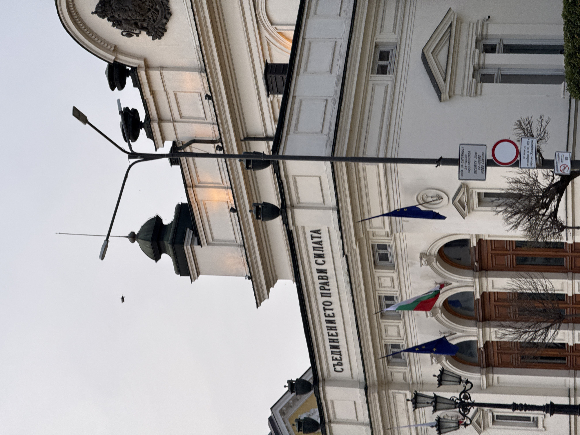 The image shows a low-angle, upward-facing shot of a building facade in Sofia, Bulgaria. The building is white and has prominent architectural details, including moldings, recessed windows, and a dome-shaped top. The phrase "СЪЕДИНЕНИЕТО ПРАВИ СИЛАТА" (Unity Makes Strength) is inscribed on the building. Bulgarian and European Union flags are mounted on the building, and there are vintage-style streetlights present. Signs that read "ЗОНА ЗА СИГУРНОСТ 24-ЧАСОВ ВИДЕОКОНТРОЛ SECURITY ZONE 24-HOURS VIDEO CONTROL" are visible, indicating surveillance in the area. Bare trees and branches are noticeable along the facade. The sky is overcast and appears to be a light grey color.