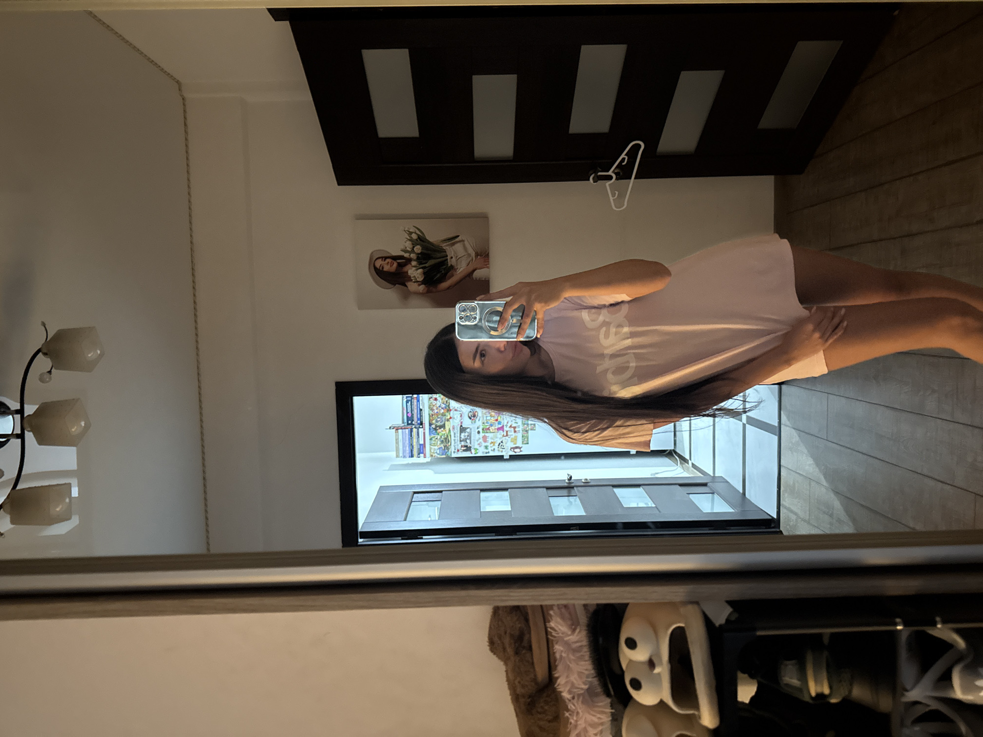 The image captures a young woman standing in front of a full-length mirror, taking a selfie with her phone. She is wearing a light pink oversized t-shirt. The mirror reflects a room with wooden floors and a closed dark wooden door with three horizontal rectangular panels. To the left of the door, a white hanger is visible. On the wall above the door, a framed picture depicts a woman holding a bouquet of tulips.
The room appears to be a bedroom or a dressing area. To the left of the mirror, a shelf unit is partially visible, holding stuffed animals and what appear to be slippers. A light fixture with three square shades hangs from the ceiling. The overall lighting suggests an indoor environment, likely a residential setting. There are no explicit indicators of the specific location within Chișinău, Moldova, beyond it being an indoor private space.