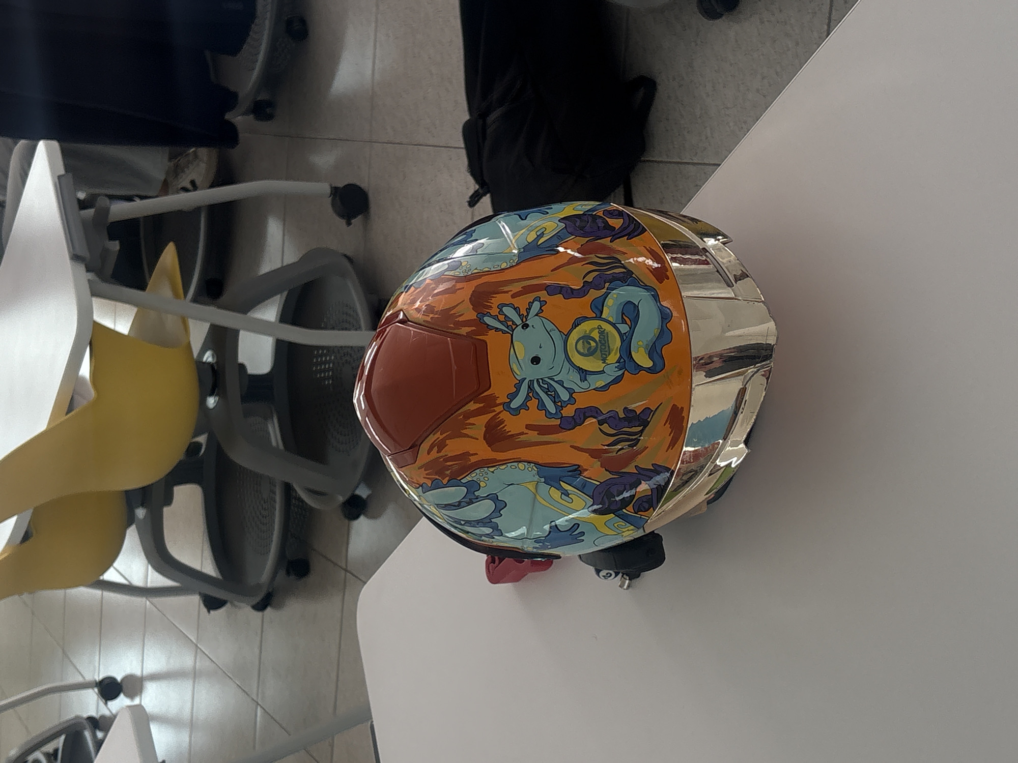 A motorcycle helmet sits on a white table in what appears to be an indoor environment. The helmet features a distinctive orange and blue design depicting cartoonish axolotls. To the left of the table are multiple chairs, some with yellow seats and others gray. In the background, a black backpack rests on a tile floor. While the city of Heroica Puebla de Zaragoza, Mexico is mentioned, there's no information in the image that directly confirms that it's located in that city. There are no people visible in the frame.
