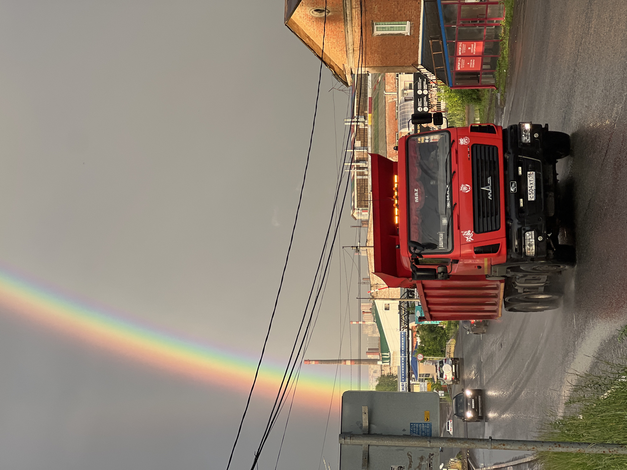 The image captures a street scene in Izhevsk, Russia, under a cloudy, possibly rainy sky, as indicated by the wet asphalt and the presence of a rainbow. The primary subject is a bright red MAZ truck, prominently positioned in the foreground, with its hood open. The truck is parked on the wet street, with water splashing from its wheels.
The setting appears to be a commercial or industrial area. Buildings with brick facades and storefronts line the street. One building has a blue awning with red signs, and another displays advertisements. In the background, industrial chimneys and other structures are visible, suggesting a factory or processing plant.
A faint rainbow arcs across the gray sky, a stark contrast to the otherwise somber weather. Several other vehicles, including cars and possibly another truck, are seen on the road, suggesting ongoing traffic. Power lines crisscross the frame, adding to the urban feel.
Visible text includes "MAZ" on the truck, along with Cyrillic text that appears to be advertising slogans or company names such as "МОТОРЛЭНД" and possibly a name like "ДЯДЯ РУСЛАН." A license plate on the truck reads "c504 XT 18."
The overall impression is a snapshot of a moment after or during a rain shower, with a truck parked on a wet street in a Russian town, accompanied by the atmospheric phenomenon of a rainbow. The image feels somewhat mundane yet picturesque due to the vibrant red truck and the natural wonder of the rainbow.