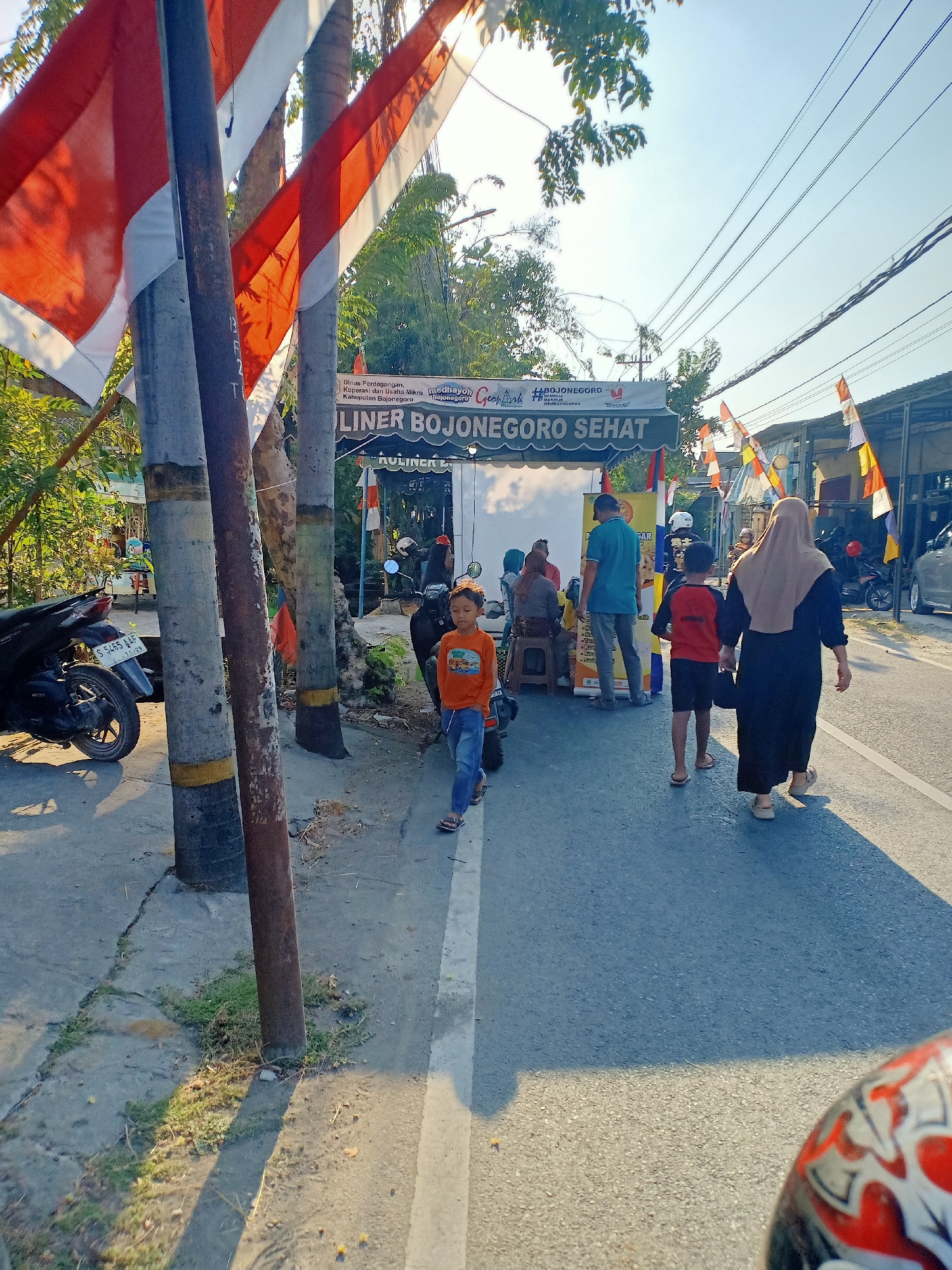 This is a medium shot photograph of a street scene. In the foreground, to the left, is a motorcycle. To the right, a young boy in an orange t-shirt and blue jeans walks towards the right of the frame. Behind him, a woman in a black dress and beige hijab walks away from the camera. Behind them, several people are gathered around a stall with a banner that reads "KULINER BOJONEGORO SEHAT". Flags are strung across the street. The background shows trees, power lines, and buildings. The sun is shining, casting shadows. The image appears to be taken in a city, possibly in Indonesia, based on the text on the banner.