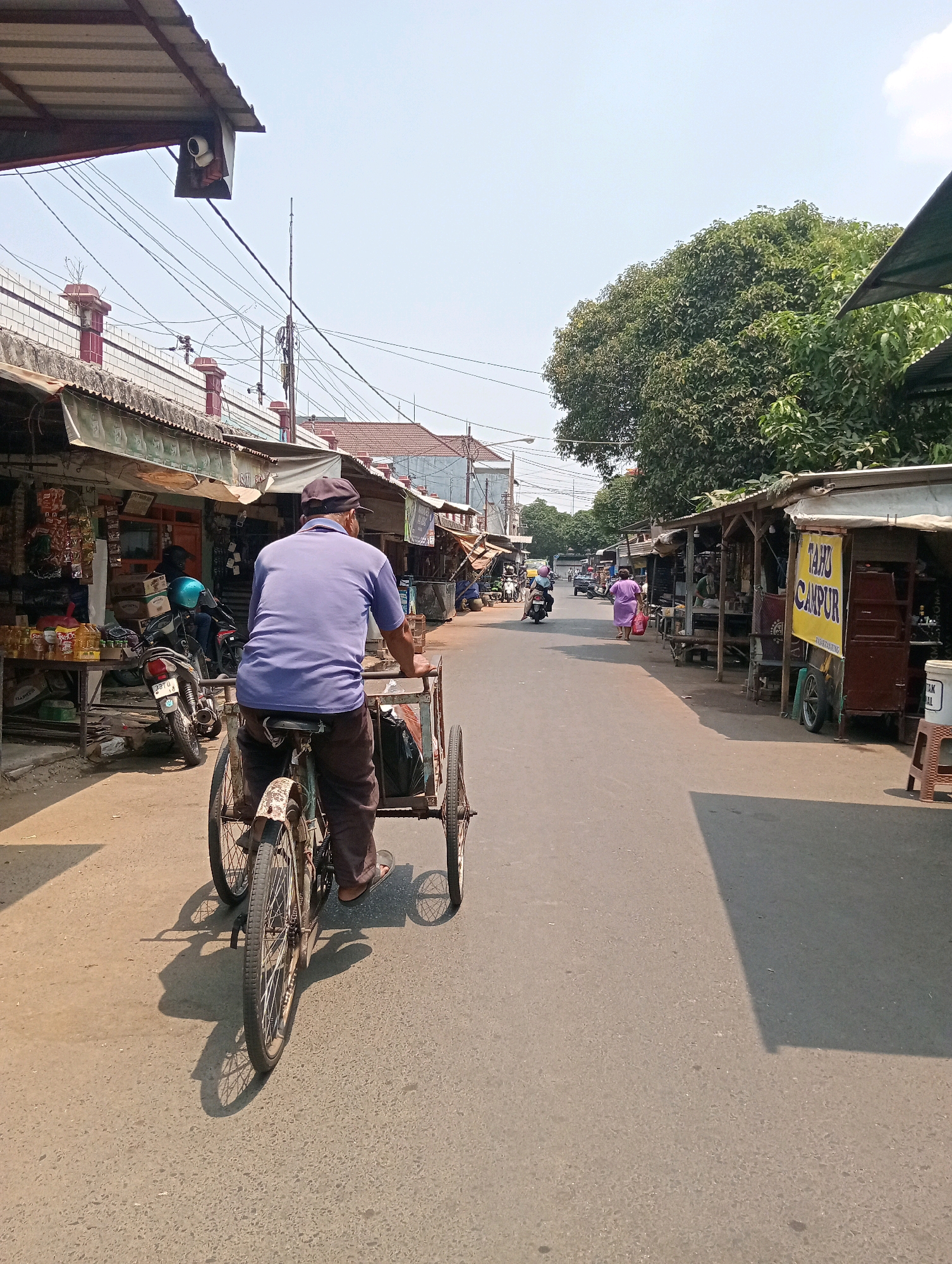 The image depicts a street scene in Jagalan, Indonesia. In the foreground, a man wearing a purple shirt and a cap rides a three-wheeled bicycle. He is moving away from the camera and is positioned on the right side of the road. The road is paved and appears to be a typical urban street.
Along the sides of the road, there are numerous small shops and market stalls, indicative of a local marketplace. These structures have corrugated metal roofs and wooden supports, with goods displayed outside. On the left side of the street, there are shops selling various items, including bottled drinks and other merchandise. A motorcycle is parked in front of one of these shops.
On the right side of the street, more market stalls are visible, some with signage. One stall has a large yellow banner with text that appears to be in Indonesian, possibly advertising food or products.
In the background, other people can be seen, including a woman in a purple dress walking with shopping bags and individuals on motorcycles. The environment is characterized by a clear sky and lush green trees are visible on the right side of the frame, contrasting with the man-made structures. Overhead power lines crisscross the scene, a common feature in many urban areas. The overall impression is of a bustling local market area on a bright, sunny day.