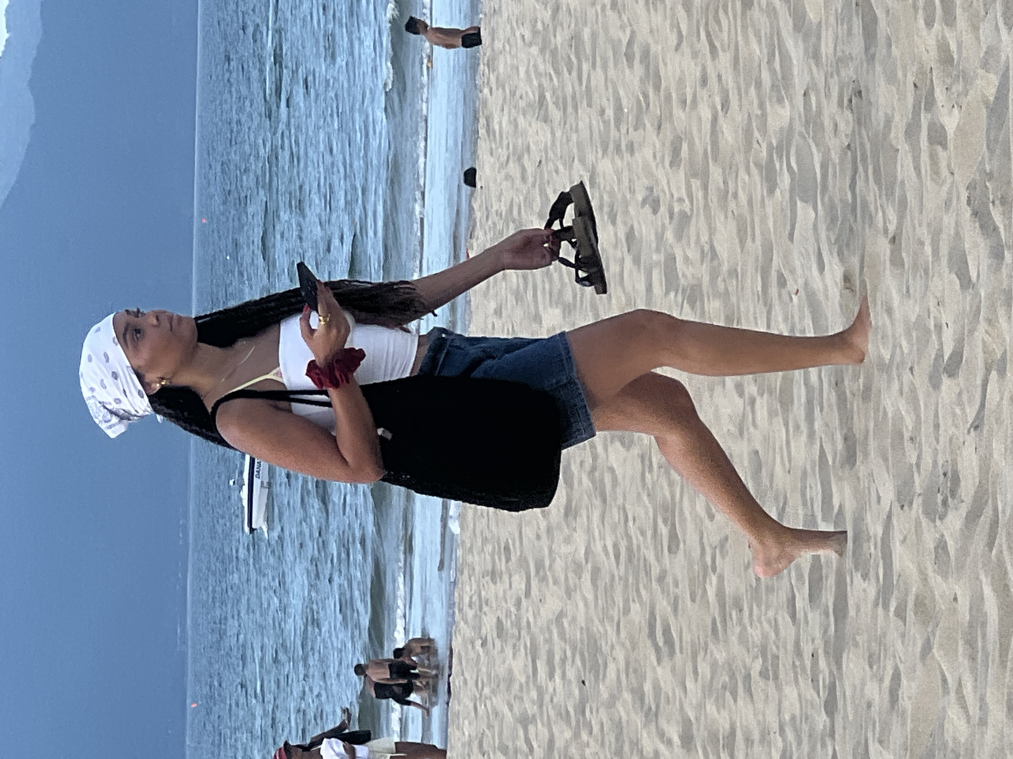 A young woman with braided hair and a white bandana walks across a sandy beach. She is wearing a white crop top and denim shorts, carrying a black bag over her shoulder and holding a mobile phone in her right hand, while her left hand clutches a pair of dark sandals. The ocean is visible in the background with a small white boat on the water. Other people are scattered on the beach and in the shallow water, suggesting a public beach setting in Đà Nẵng, Vietnam. The sky is overcast and the overall atmosphere is serene.