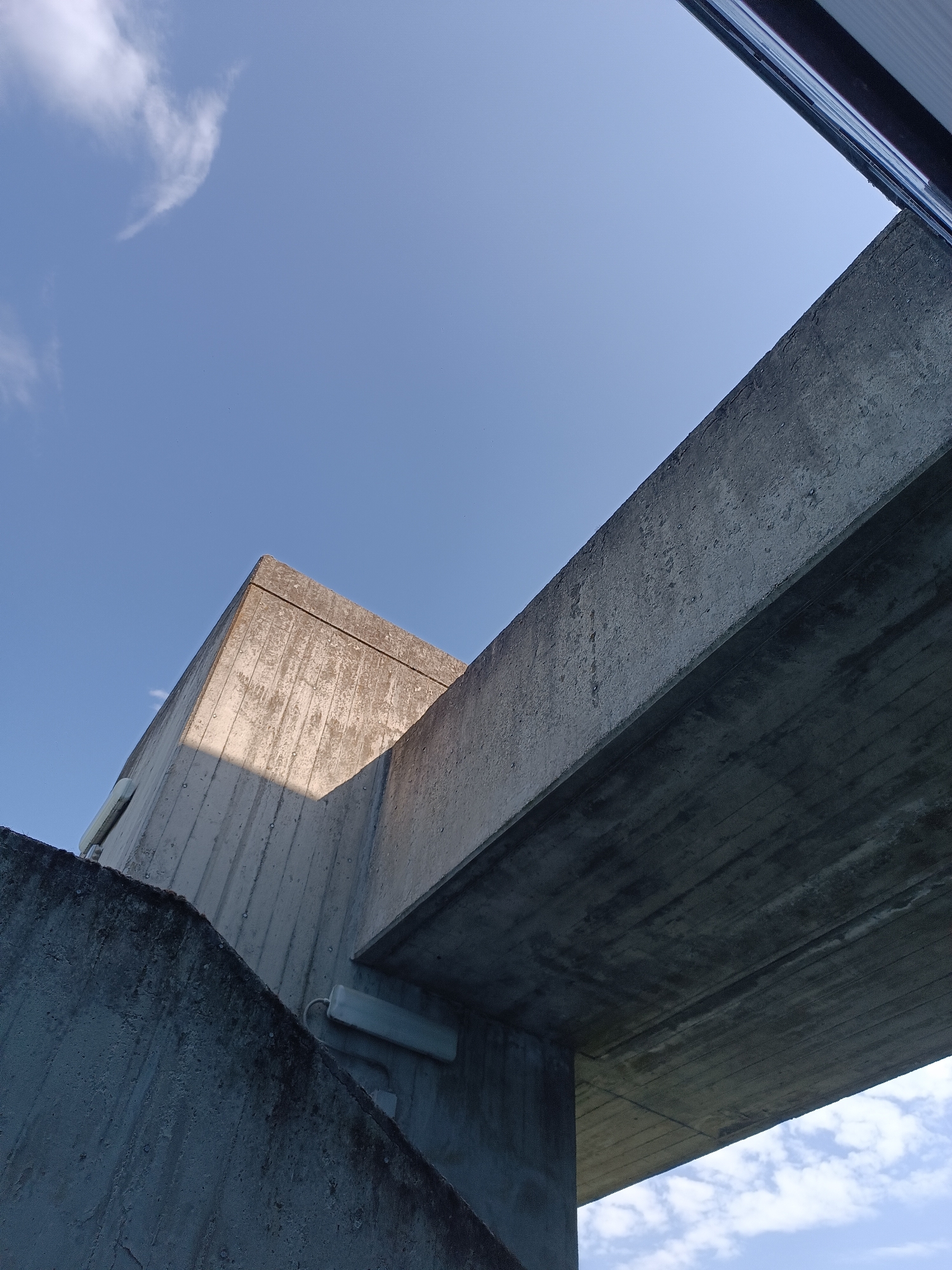 The image captures a low-angle perspective of concrete structures against a clear blue sky with scattered white clouds. The architecture appears modern and geometric, with sharp edges and a rough concrete texture. Sunlight casts a distinct shadow across the corner of one of the structures, highlighting its form and the contrast between light and shade. There are no people or visible signs of activity. The environment suggests an outdoor urban setting, possibly a park, bridge, or modern building complex. The image does not provide any specific indicators of its location in Casalmaggiore, Italy.