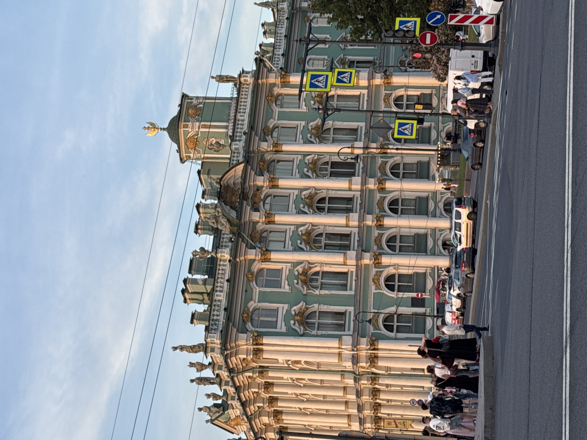 This image depicts the Winter Palace in Saint Petersburg, Russia, bathed in the warm light of a setting sun. The grand Baroque building, painted in a pale green with white and gold accents, dominates the frame. Its facade is adorned with numerous columns, arched windows, and ornate sculptures. Atop one of the turrets, a golden double-headed eagle emblem shines.
In the foreground, a wide asphalt street with white lane markings stretches across the bottom of the image. Pedestrian crossings are visible, indicated by yellow diamond-shaped signs featuring a pedestrian silhouette. Traffic lights and street signs are mounted on metal poles.
On the sidewalk in front of the palace, a group of people are gathered. Some are standing, others appear to be walking. They are dressed in casual summer attire. A few cars, including a white van and a dark-colored sedan, are parked or driving on the street. The sky above is a clear, soft blue with wispy clouds. The overall atmosphere is one of urban activity against the backdrop of historical grandeur.