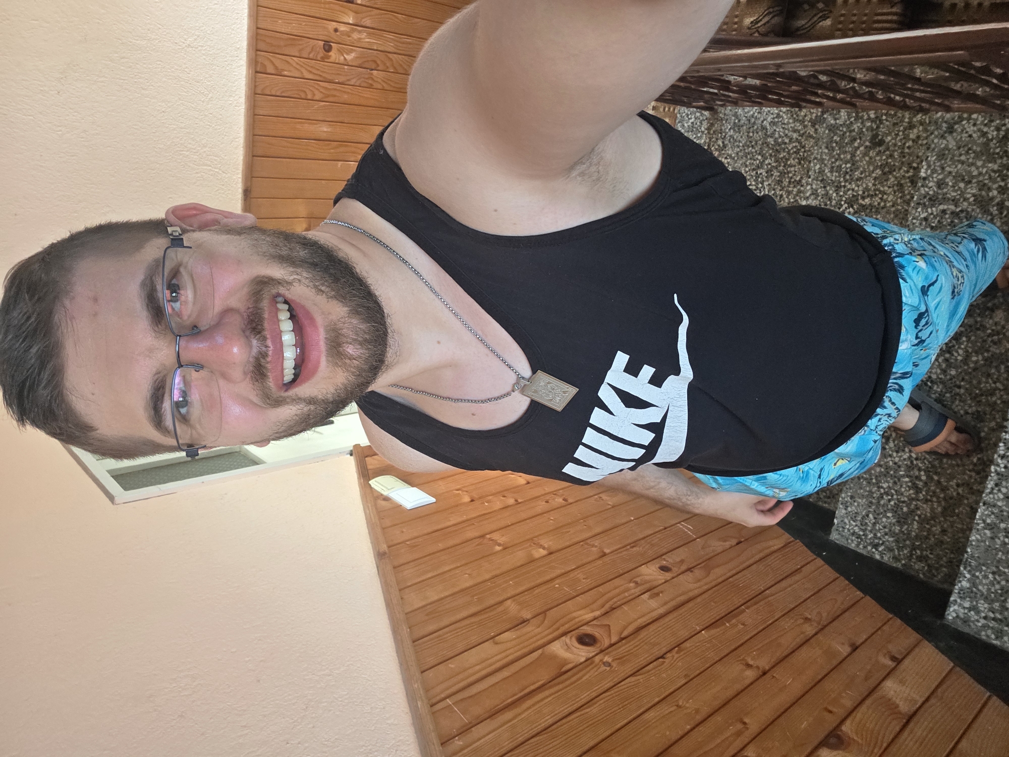 A man with a beard and glasses is standing in a room in Yagodovo, Bulgaria. He is wearing a black Nike tank top and blue patterned shorts. He has a silver chain with a pendant. The room has a wooden wall and a tiled floor.