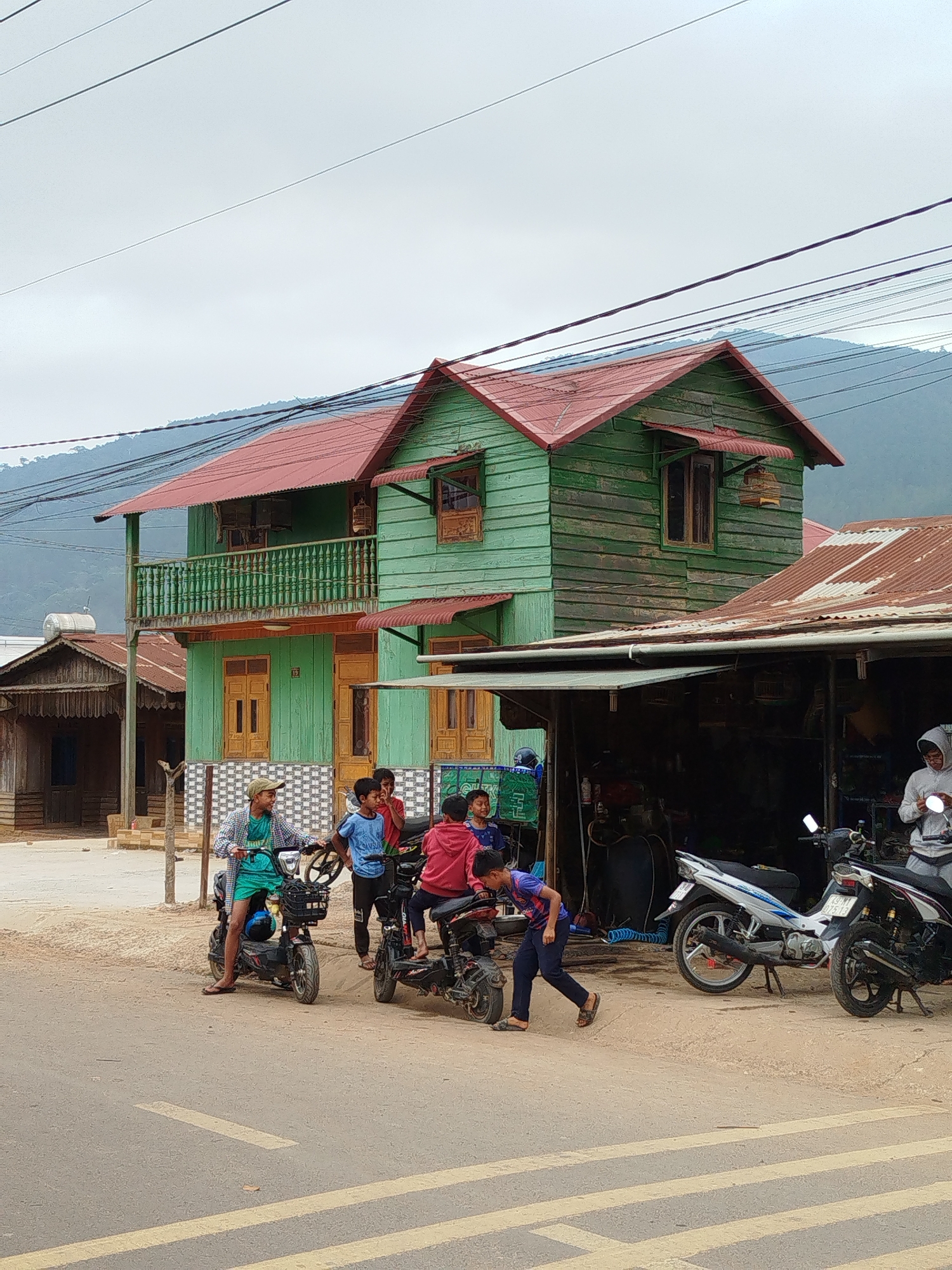 The image captures a street scene in what is likely Đarahoa, Vietnam. A two-story wooden building with green siding and a red roof stands prominently. Adjacent to it is a smaller building with a corrugated metal roof, under which are parked motorbikes. Several young people are near the motorbikes. One is on a motorbike with a basket, one on another motorbike, and one walking, seemingly looking at or interacting with the motorbikes. Overhead, power lines are visible against a cloudy sky. A mountain range is discernible in the background.