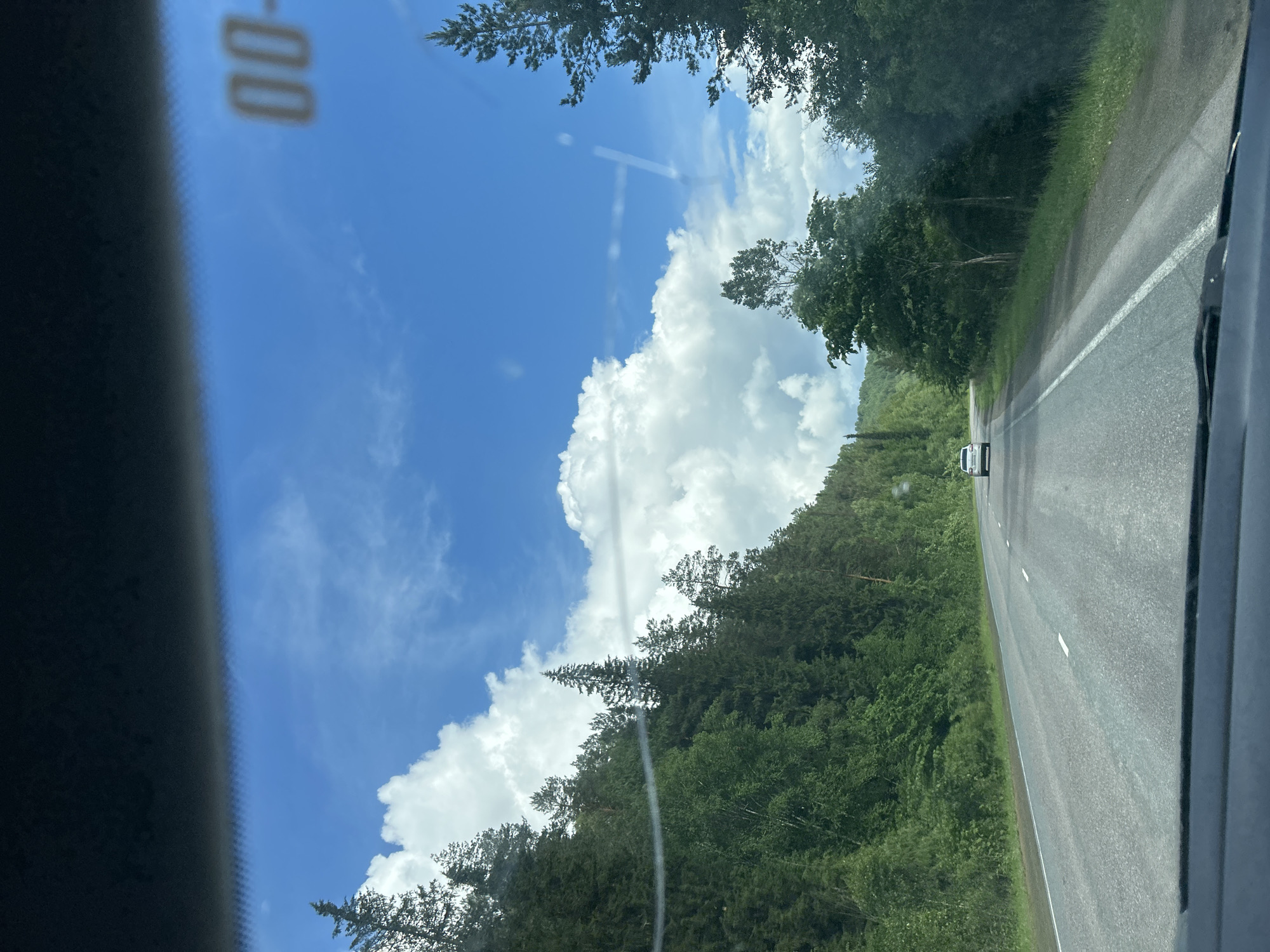 A white car drives down a paved road through a forest under a bright blue sky with fluffy white clouds. The road is lined with trees and vegetation. The image appears to have been taken from inside a vehicle, possibly through the windshield. There is a dark dashboard visible at the bottom of the frame. In the upper right corner of the image, there is a blurred marking that appears to be "00-" on the windshield. The specific location in the city of Усмангали, Russia is not identifiable from the visual information provided.