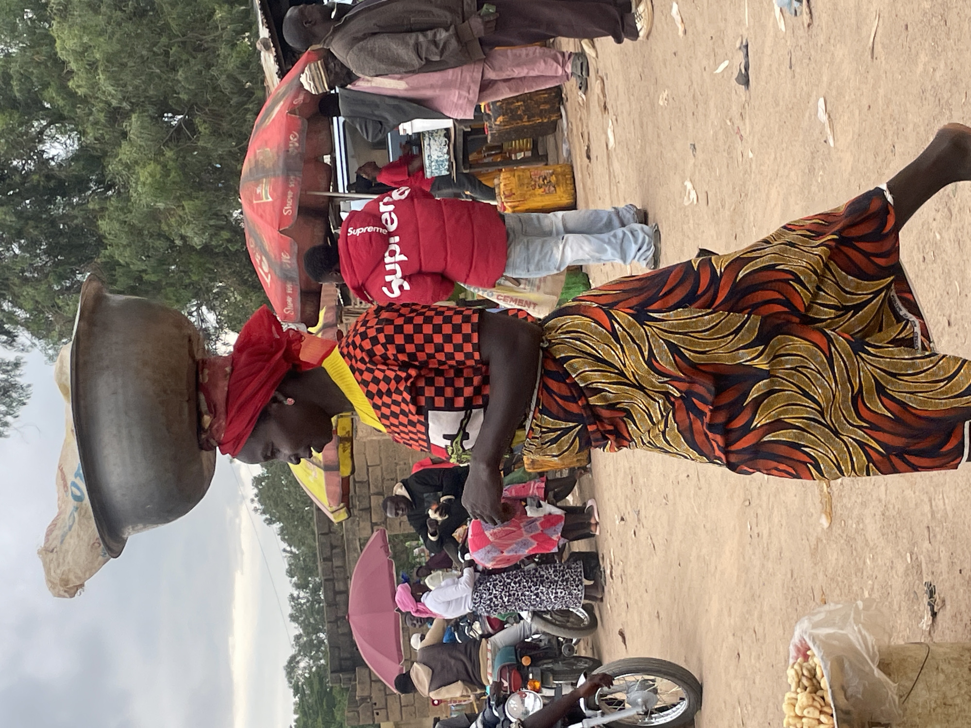 The image depicts a street scene in Wang, Nigeria, possibly a market. A woman in the foreground, wearing a red headscarf, a black and orange checkered shirt, and a patterned skirt, carries a large metal bowl filled with goods on her head. She is walking towards the right side of the frame. In the background, several people are visible, some under red and yellow umbrellas. There are motorcycles, yellow fuel cans, and what appear to be market stalls. The ground is dusty earth, and the overall environment suggests a bustling outdoor setting.