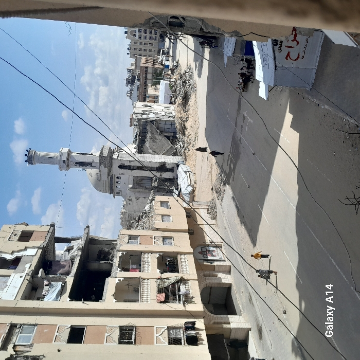 The image displays a street scene in Khan Yunis, Palestinian Territory, characterized by extensive structural damage to buildings. A paved road with a faded white center line occupies the foreground, strewn with scattered dirt and debris. Three children are walking on the left side of the street. To the immediate left, a multi-story building shows significant damage across multiple floors, with exposed interiors, broken windows, and missing sections; clothing hangs from several balconies.
In the midground, a prominent mosque with a large dome and a tall, slender minaret stands amidst considerable rubble and a partially collapsed white tent-like structure. Two adults are seen walking on the street in the direction of the mosque. Damaged multi-story buildings also line the right side of the street, where a group of approximately 5-7 individuals are seated around a low table under two white tarpaulin shelters. One shelter bears partial Arabic text, including "حراج" (haraj). The background shows additional urban structures, some appearing damaged. The sky is partly cloudy with patches of blue. Numerous black power lines or cables traverse the upper portion of the scene.