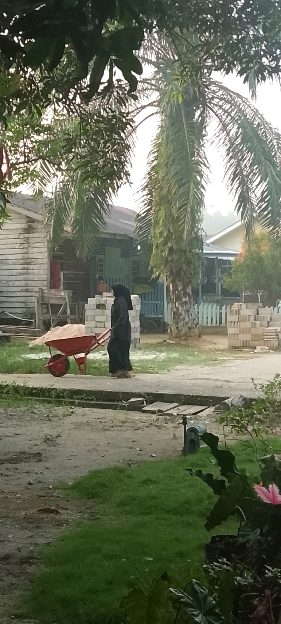 A woman wearing a black hijab and long dark clothing walks to the right, pushing a red wheelbarrow filled with light-colored sand. She is on a paved surface in front of a stack of pale bricks. To her left is a wooden structure with a white-sided building behind it. Palm trees are visible in the background and foliage is present in the foreground. The scene appears to be in a residential or semi-rural area. The light suggests it is daytime. The location is identified as Harapan Jaya, Indonesia.