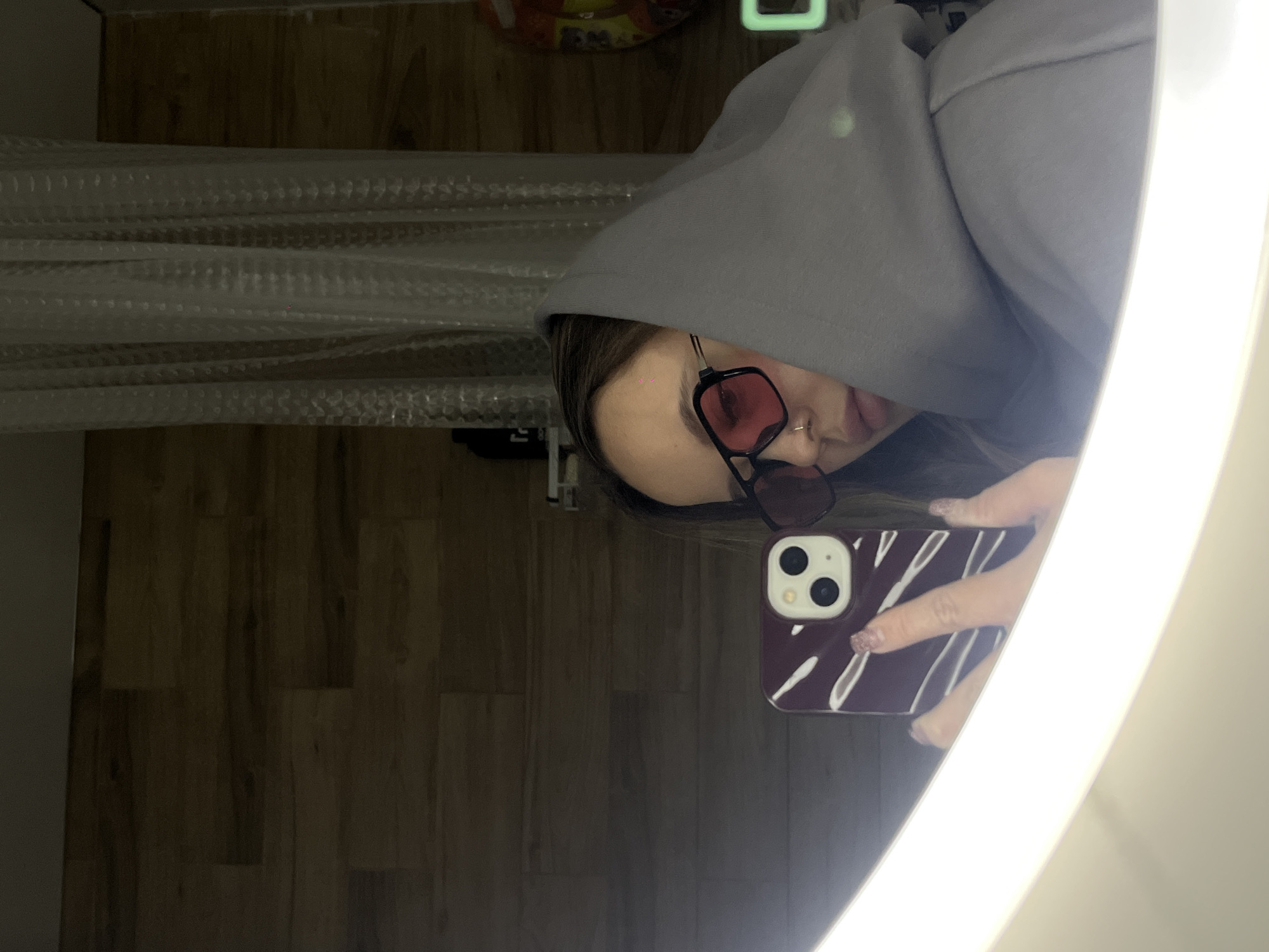 This image is a mirror selfie taken by an individual in an indoor setting, likely in Krasnodar, Russia. The photo is rotated 90 degrees clockwise.
The central subject is a person, partially obscured by the hood of a grey hooded sweatshirt. They are wearing dark-framed sunglasses with reddish-pink tinted lenses, which cover their eyes. A small nose ring is visible. The person is holding a smartphone in their right hand to capture the reflection. The smartphone has a distinct dark purple or maroon case with white, abstract, possibly leaf-like patterns, and its dual camera system is clearly visible on the back. The person's nails have a light-colored, possibly glittery, polish.
The selfie is captured in a brightly lit mirror, which features a prominent LED light strip along its edge, creating a strong glow around the reflection.
The background visible in the mirror's reflection includes dark brown wooden panels, suggesting either a floor or a wall. To the left of the frame, there is a translucent, textured curtain or screen, possibly a shower curtain or window blind, characterized by a pattern of small bumps or ridges. Higher up in the background, a blurry object resembling food packaging and a distinct green-glowing rectangular light (possibly from an electronic device) are visible.
The image depicts a casual moment of self-photography. No discernible text is visible within the frame.