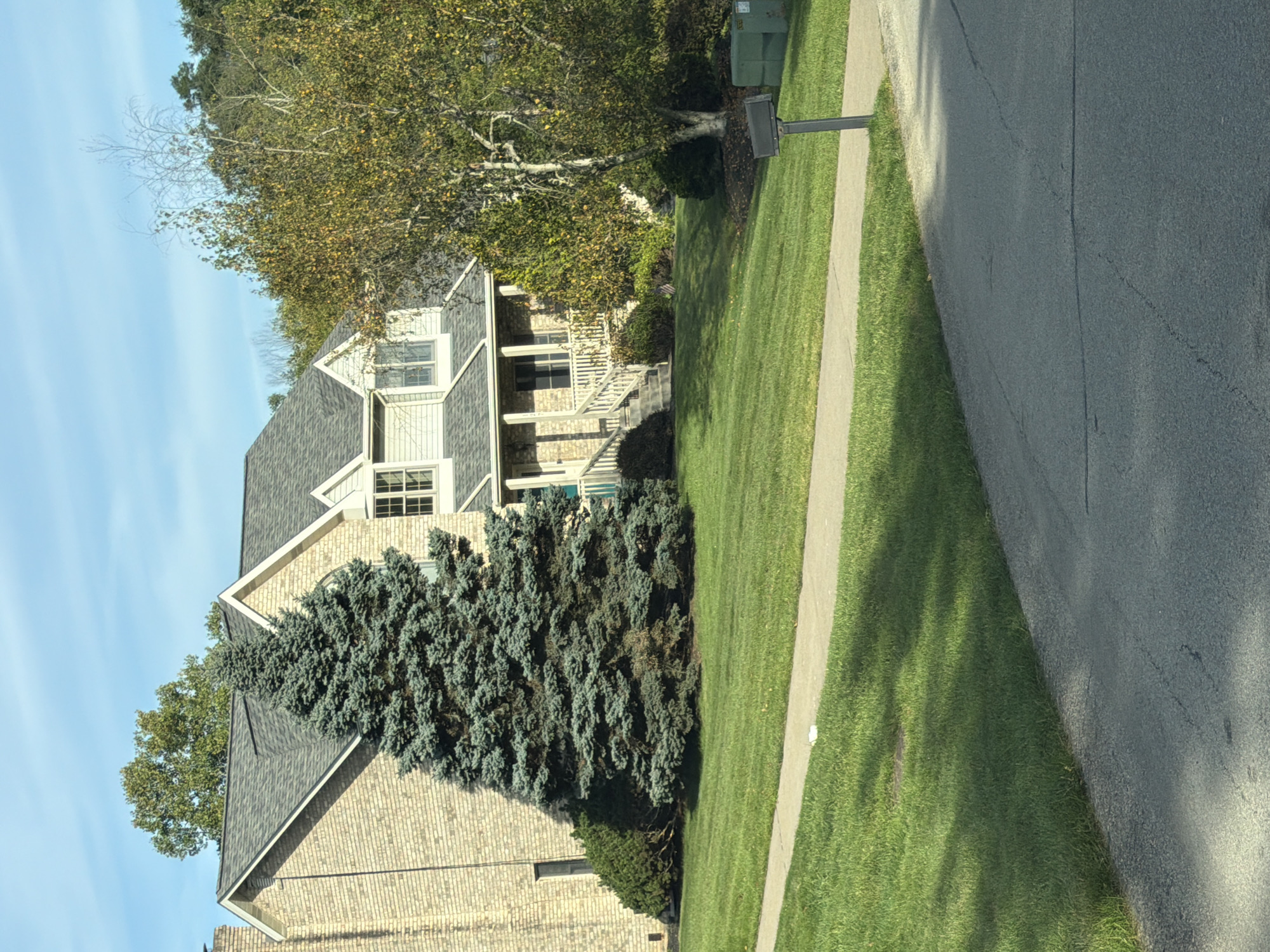The image depicts a suburban scene with a two-story brick house situated on a gently sloping, well-maintained lawn. A paved driveway curves into the foreground, and a concrete sidewalk runs horizontally across the middle ground. To the left of the house, a large, mature evergreen tree dominates the landscape. To the right of the house, a deciduous tree with yellowing leaves suggests an autumn setting. A mailbox stands near the edge of the driveway on the right side of the frame, next to two large green bins. The sky is a clear blue with wispy clouds. The house features a dark gray roof, light-colored siding on the upper level, and white trim around the windows and porch. The front entrance is accessed by a set of stairs leading to a covered porch. The overall impression is one of a quiet, residential neighborhood.
