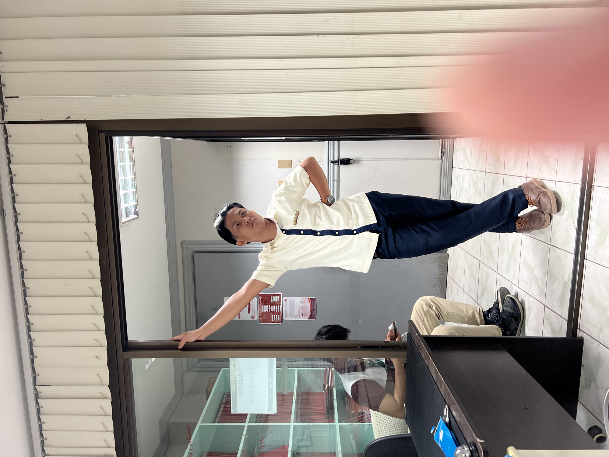 The image shows a man leaning against a window frame, appearing to be in an indoor setting that resembles an office or a school hallway. He is wearing a light-colored polo shirt with dark trim and buttons, dark trousers, and brown shoes. His left arm is extended, resting on the window frame, and his right hand is on his hip. He is looking towards the camera with a neutral expression.
In the background, another person is visible, seated with their legs up. They are wearing khaki pants and dark shoes. Behind them, shelves filled with what appear to be binders or files are visible. The walls are tiled in a light color. A door is visible in the distance.
The scene is indoors, and the lighting suggests it could be daytime, although no specific cues for time of day are apparent. The overall atmosphere seems casual and everyday.
There is some text visible on a sign inside the glass of the window, which reads "CAUTION GLASS DOOR" in reverse, indicating it is seen from the outside. Another sign on the wall behind the main subject appears to have some text and graphics, but it is too blurry to be read.
The image is oriented vertically, with the subjects appearing to be standing upright relative to the actual scene, but rotated in the image file itself.