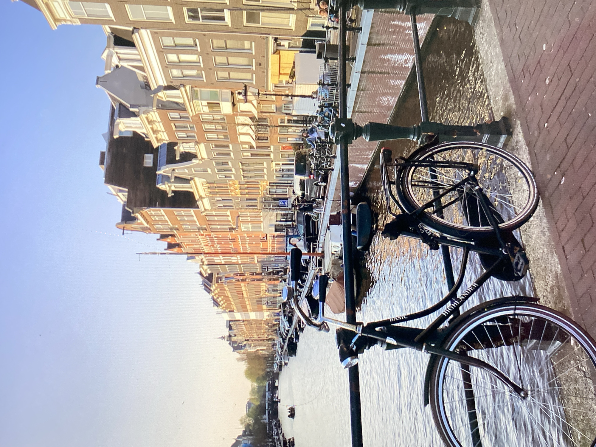A black bicycle is parked on a brick walkway next to a canal in Amsterdam, Netherlands. The bicycle has a basket on the rear and a dynamo light on the front. The canal is lined with narrow, tall buildings, many of which have gabled roofs. Several boats are visible in the canal, and a few people are walking and sitting along the canal bank. The sun is setting, casting a warm glow on the buildings. The sky is clear and blue.