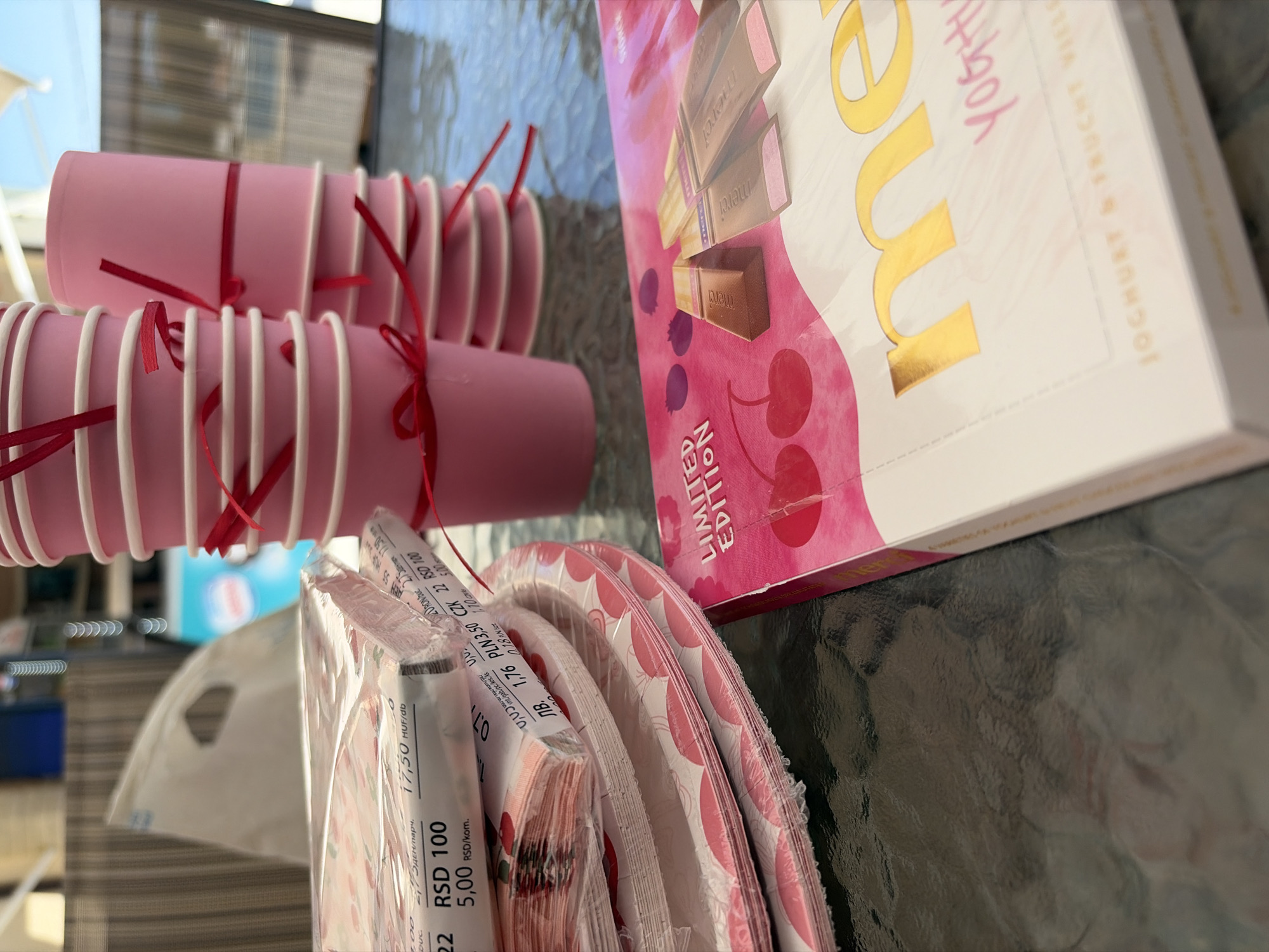 The image shows a table with party supplies. In the foreground, there is a pink box of Merci chocolates with the "Yoghurt & Frucht Viel" flavor. To the left of the chocolates, there are stacks of pink paper plates wrapped in plastic and a stack of folded pink napkins, also wrapped in plastic. Behind the plates and napkins, two stacks of pink paper cups tied with red ribbon are visible. The cups are sitting on a glass-topped table. In the background, there is a blurry outdoor setting with a striped awning and some greenery. The scene appears to be set up for an outdoor party. The location of Kolarovo, Bulgaria is not discernible from the image itself.