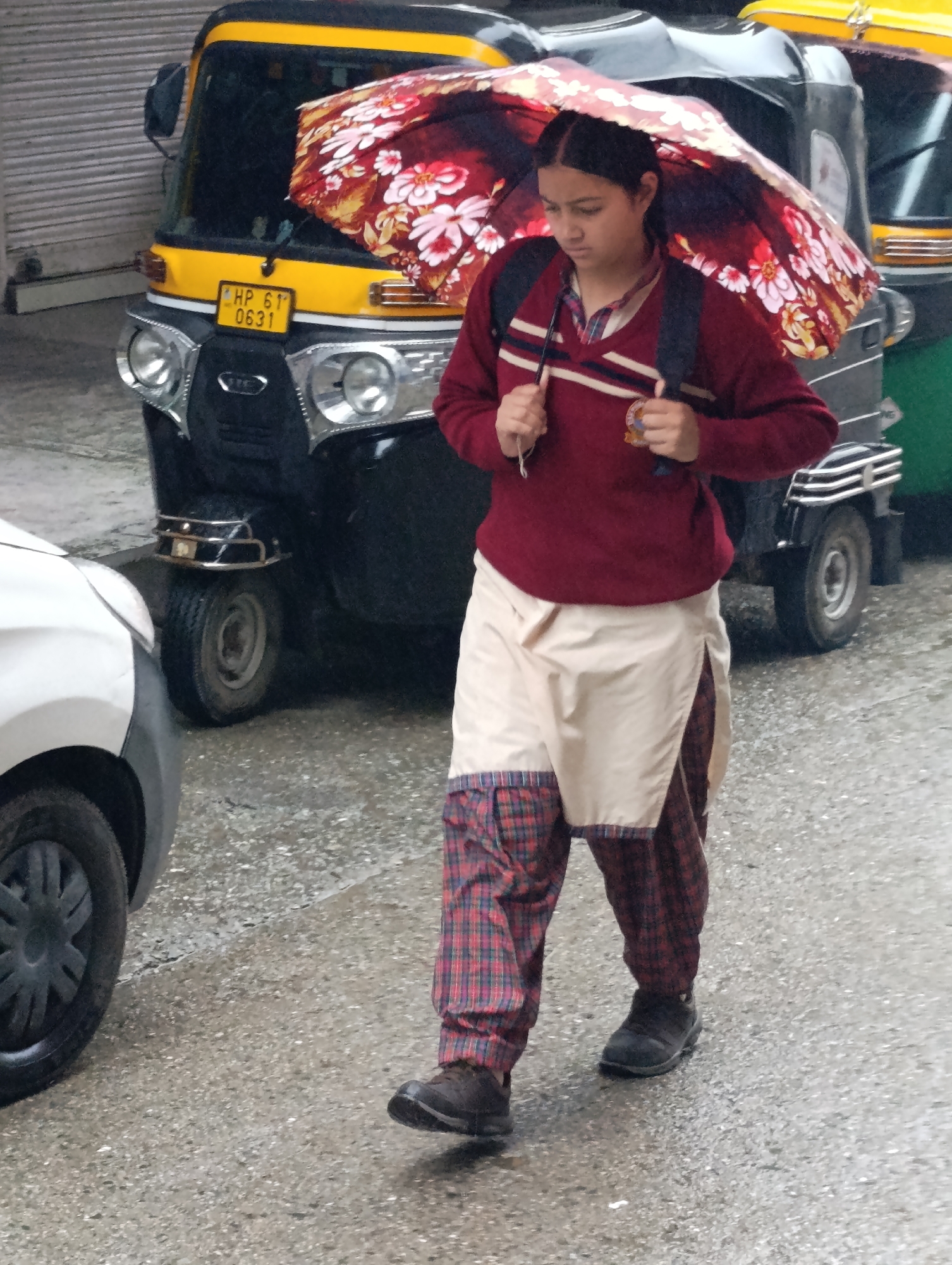 In Vashisht, India, a young girl walks under a floral umbrella on a wet, gray pavement. She wears a maroon sweater with a striped collar over a plaid shirt, and cream-colored pants with plaid accents at the bottom. She carries a dark backpack. Behind her, a yellow and black auto-rickshaw is parked, its headlights illuminated. To her left, the front of a white vehicle is visible, with its wheel turned. The ground is slick and appears to be reflecting the surroundings. The overall atmosphere suggests a rainy day in an urban setting.