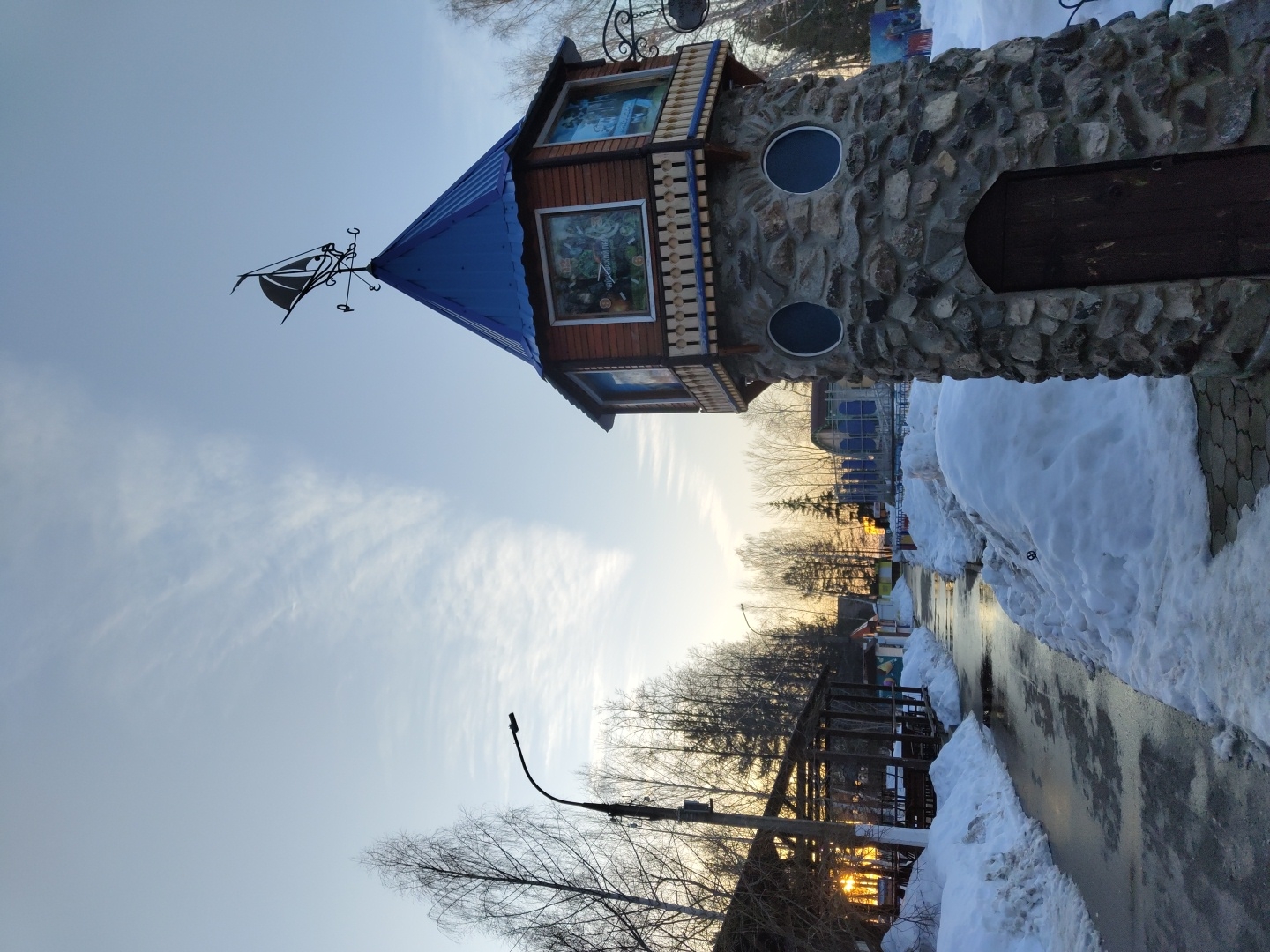 The image shows a structure resembling a small castle tower in the city of Chaikovsky, Russia. The tower has a blue, pointed roof with a weather vane on top. Below the roof, there are windows with what appear to be pictures inside. The main body of the tower is constructed from rough stones and features two round windows and a small arched wooden door. The tower stands beside a pathway partially covered with melting snow. A street light is visible in the background along with bare trees and some buildings. The sky is a light blue color with wispy clouds. The image is taken from a low angle.