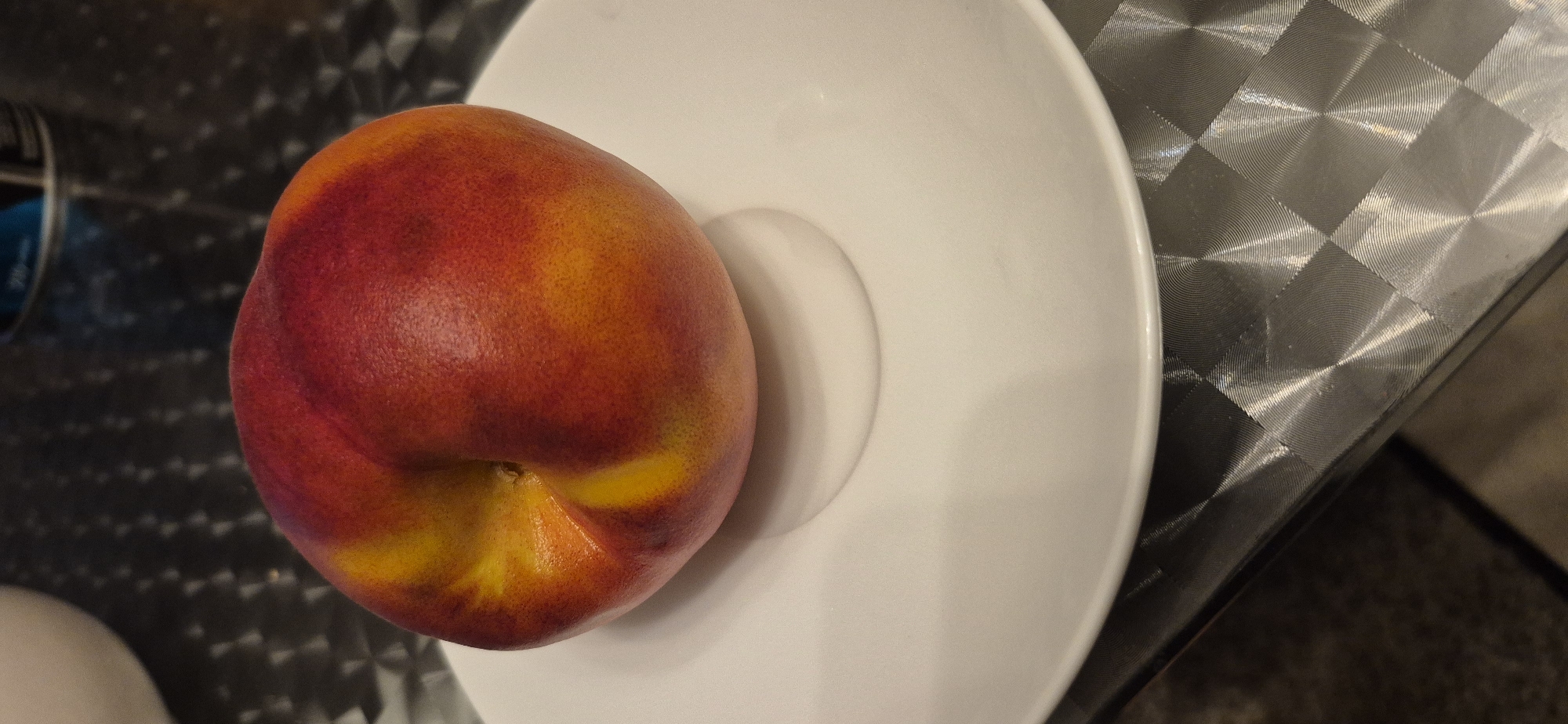 A close-up, high-angle shot shows a single, ripe nectarine with a deep red and yellow blush, resting on a white saucer. The saucer is placed on a textured, metallic surface with a diamond-like pattern, possibly a table. In the background, a blurred blue and silver cylindrical object is partially visible, and to the left, a blurry white object, possibly a bowl or another piece of dishware, is also seen. The lighting is somewhat dim, highlighting the textures and colors of the nectarine and the surfaces. The scene is devoid of any human presence or activity, and it is impossible to determine its location within the city of Baden-Baden, Germany, or any other specific city.