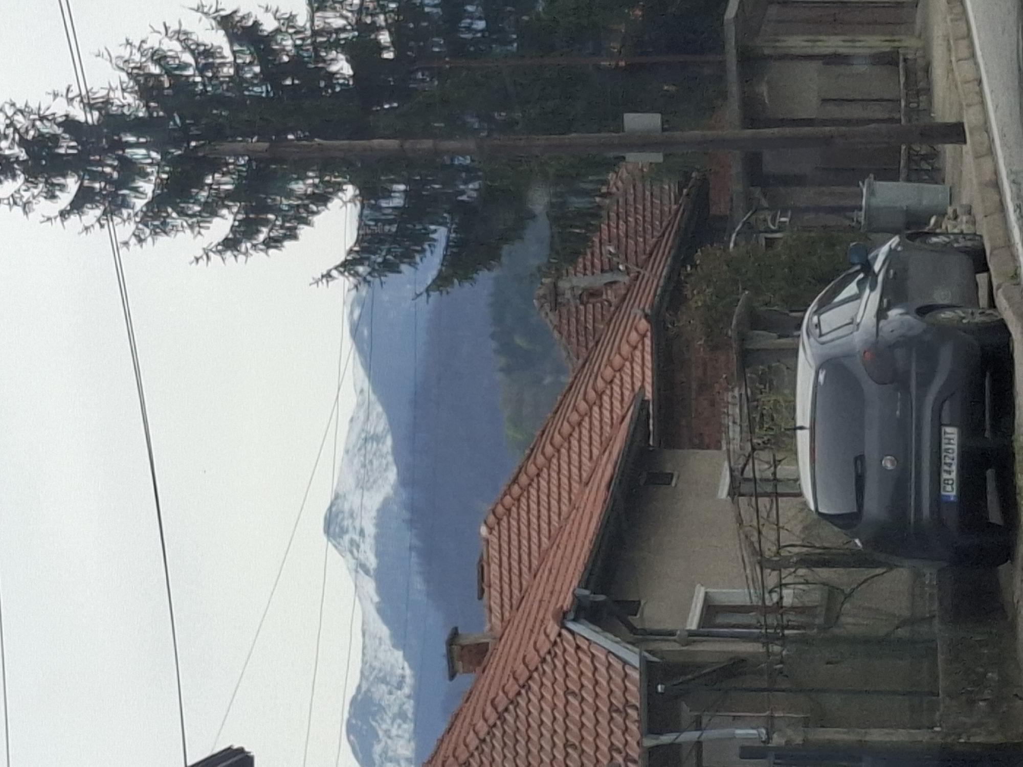 A landscape photograph captures a mountainous vista partially obscured by foreground elements, specifically a residential scene. The mountain, shrouded in a blue haze and capped with patches of white, dominates the distant background. In the mid-ground, a building is visible with a red tile roof, likely a house, indicative of a settled human environment. A tree with dense foliage frames the scene from above, partially obstructing the view of the mountain.
To the right of the frame, a dark grey car is parked adjacent to the building. The license plate, clearly visible, reads "CB 4428 HT", identifying the vehicle within a specific jurisdiction. The urban setting is further suggested by the presence of utility poles and wires that cut across the sky.
The image is taken in Barziya, Bulgaria, as evidenced by the license plate details. The scene depicts a blend of natural landscape and residential life. There are no people visible in the scene. Interactions and actions are absent as the photograph presents a static moment in time.