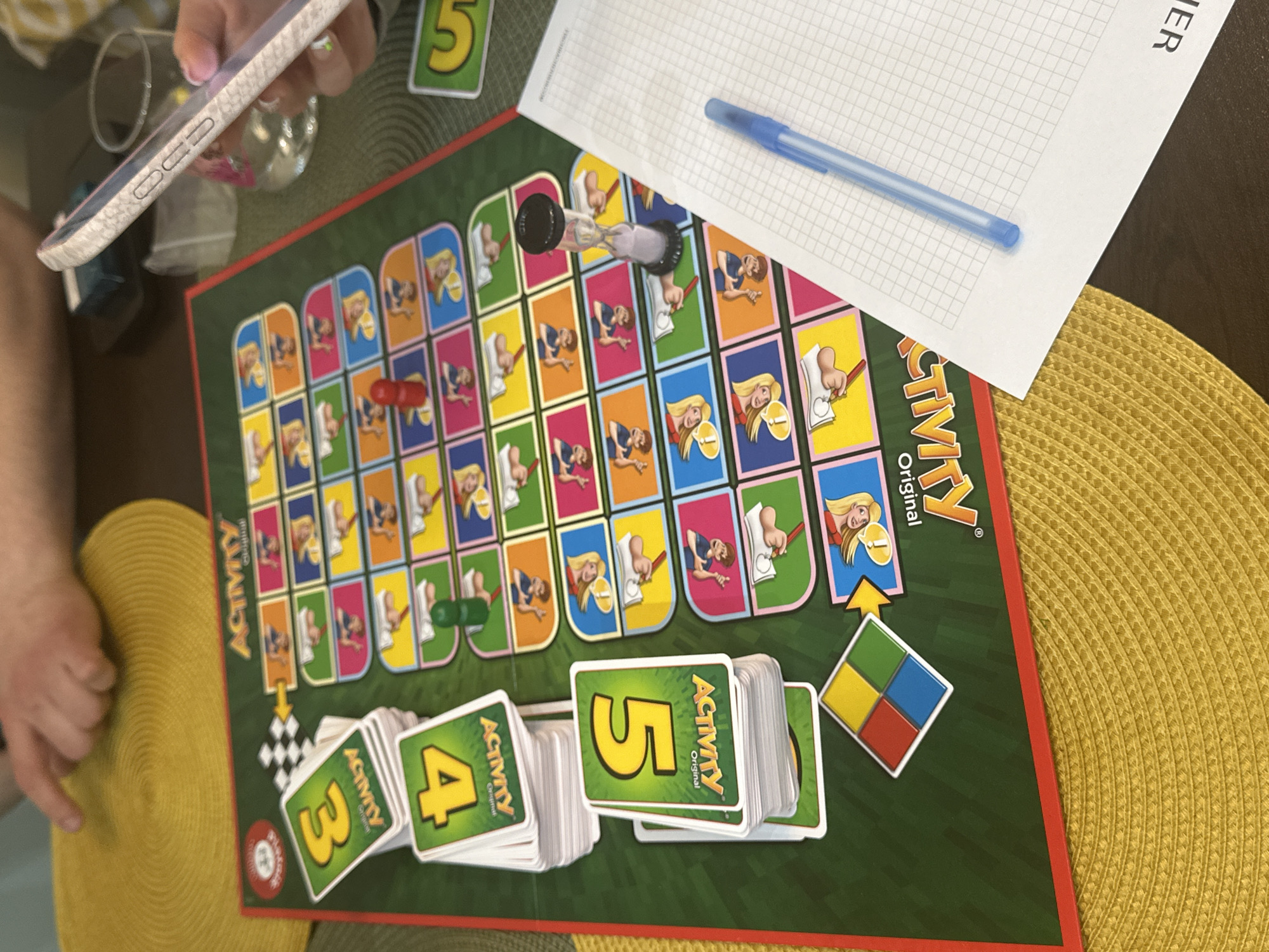 The image depicts a partially visible person holding a smartphone, alongside an "Activity Original" board game spread out on a table with a yellow placemat. The game includes playing cards with numbers 3, 4, and 5 visible, game pieces, and an hourglass timer. A notepad and a blue pen are adjacent to the game. There is no information in the image to determine the location in the city of Sofia, Bulgaria.