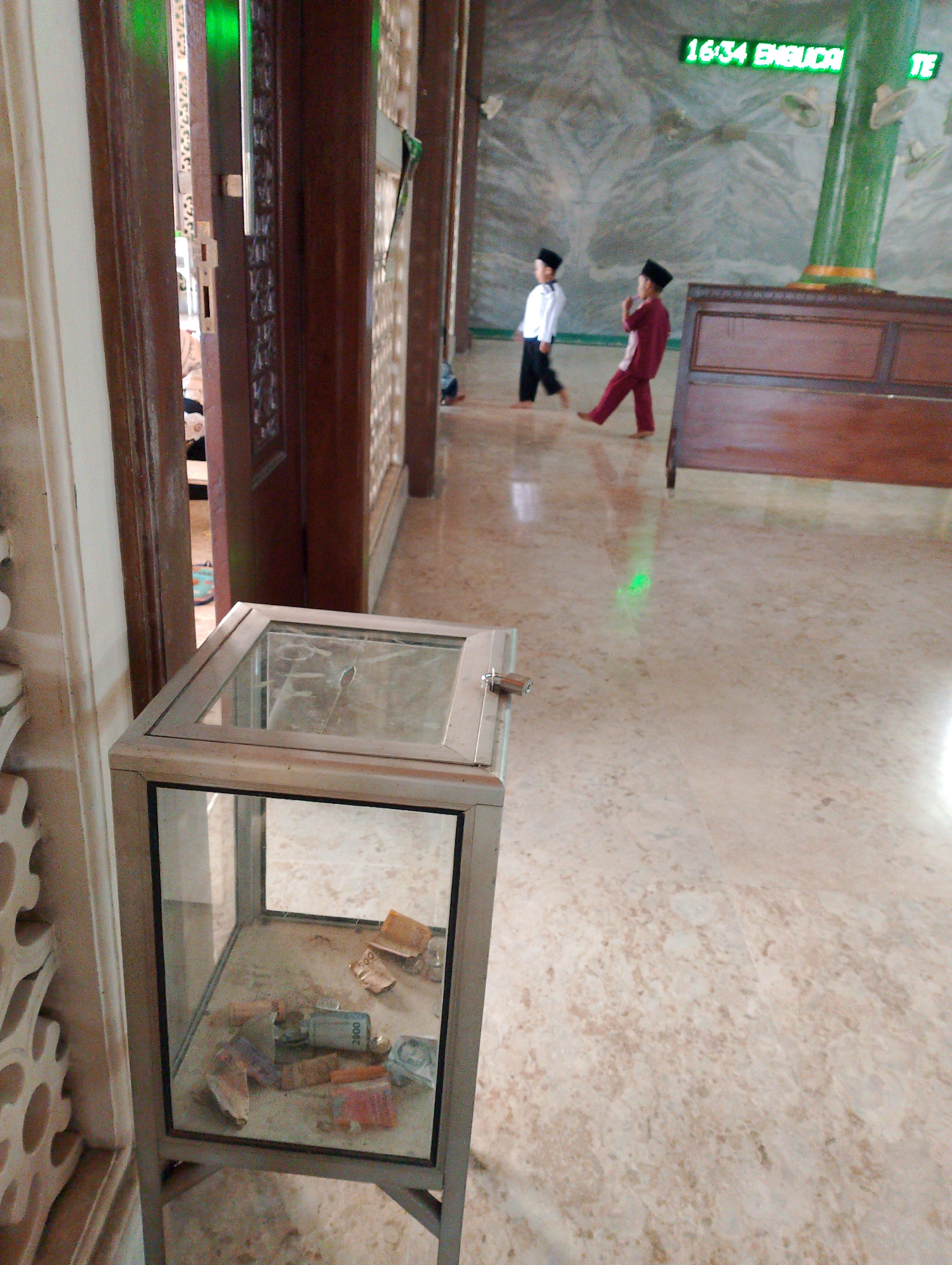 In Sukodono, Indonesia, a glass-topped donation box sits on a metal stand on a marble floor, containing several banknotes. Two young boys, one in a white shirt and black pants, the other in a red outfit, walk across the floor in the background. A digital display on the wall shows "16:34 ENGUDI TE". A wooden bench is positioned against the wall. The scene appears to be within a mosque or similar religious building, indicated by the architectural style and the attire of the children.