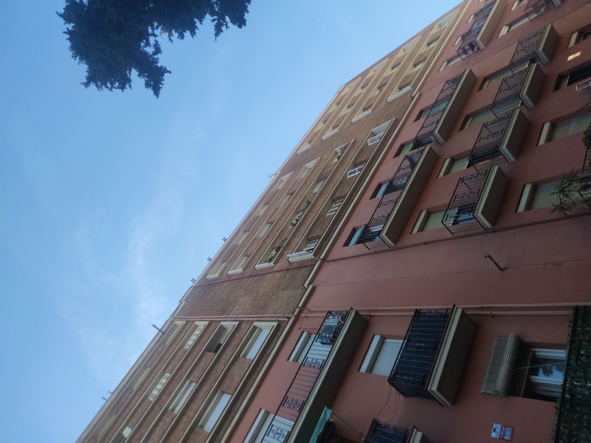 The image shows a tall building with a mix of brick and painted exterior. The building features rows of windows and balconies, some with ornate iron railings. A tree is visible in the upper left corner. The sky is blue with some light clouds. The location is Unknown, Unknown.