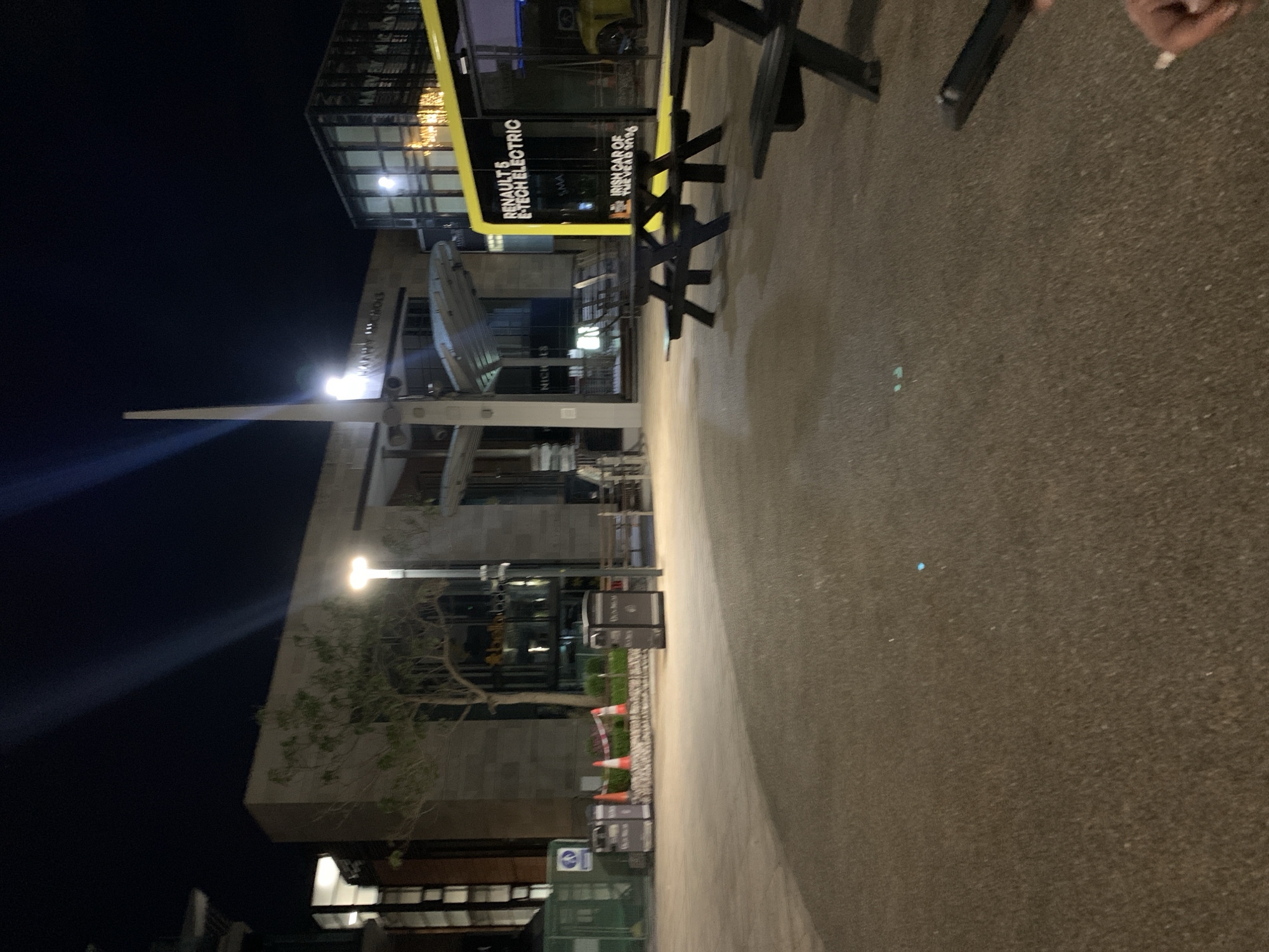 The image depicts a nighttime scene on a paved pedestrian area in Dublin 16, Ireland. A modern building with glass and concrete architecture is visible, featuring signage with "RENAULT & E-TECH ELECTRIC" and "IRISH CAR OF THE YEAR 2026" printed on it. There is outdoor seating with black picnic-style tables and benches. Construction cones are lined up along the side of the building. A tall metal structure resembling a spire is partially visible. A person's hand holding an object is in the top right corner of the image.