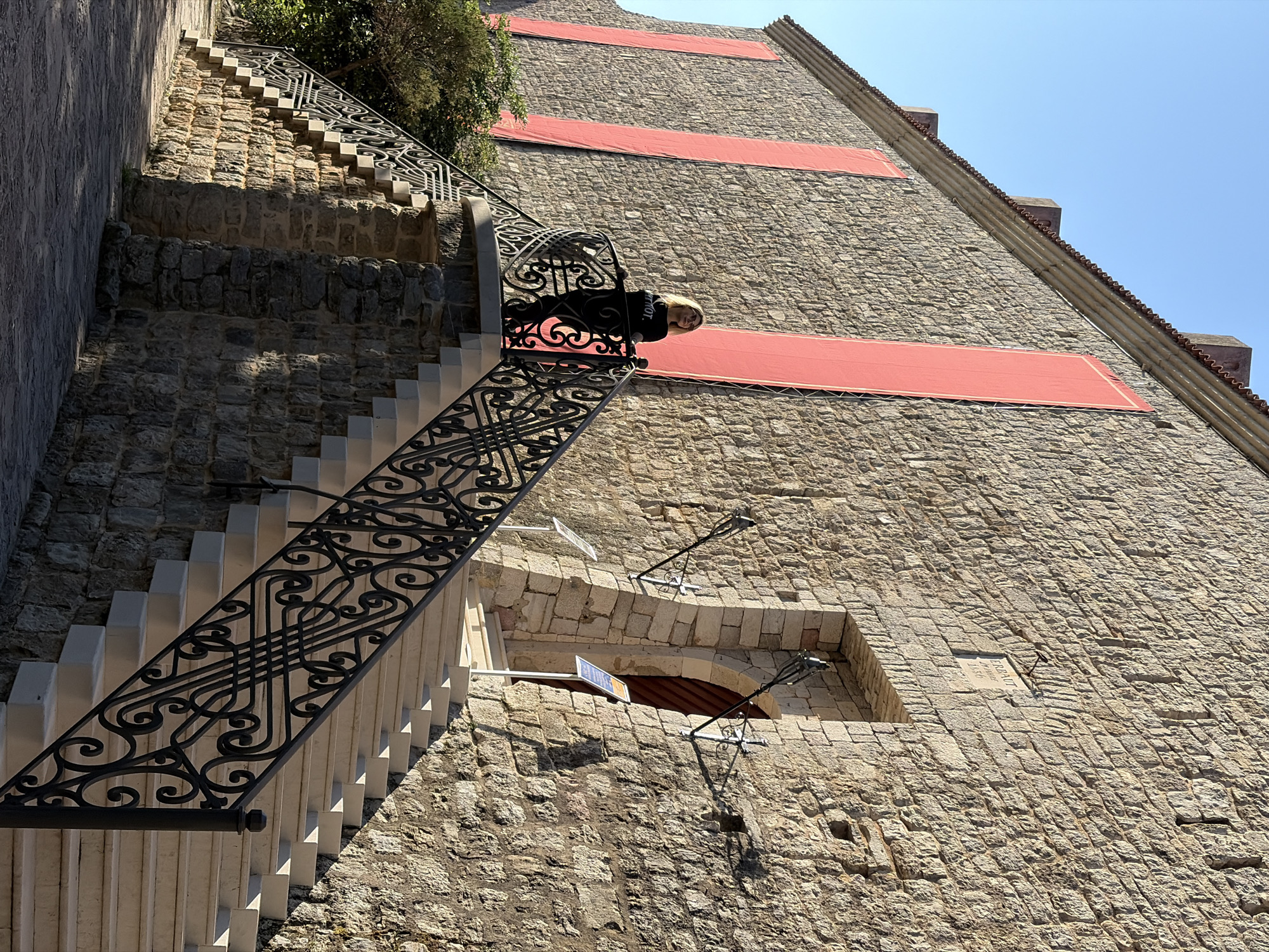 A photograph captures a grand stone wall and an ornate staircase under a bright, clear sky in Budva, Montenegro. The image is angled, making the staircase appear to ascend steeply alongside the towering stone structure. The staircase features white steps with a black, intricately patterned wrought-iron railing that twists and curves with decorative scrollwork.
A person, a woman with blonde hair wearing a black top, is visible leaning over the railing at a landing, looking outwards. The black top has the word "RIOT." printed in white text across the chest.
The massive wall is constructed from rough-hewn, light-colored stone blocks. Several red rectangular banners or awnings are affixed to the wall at regular intervals, adding a contrasting pop of color. Towards the lower section of the wall, an arched window or opening is visible, revealing a wooden door within. Two decorative, dark metal light fixtures protrude from the wall near this opening. A small stone plaque or sign is embedded in the wall to the right of this opening, which appears to read "DRAGUTIN" and "1887" beneath it, with possibly "RENOVAT" above. Lush green foliage can be seen at the top of the wall on the left side, suggesting greenery above or behind the structure. The strong sunlight casts distinct shadows, indicating a sunny daytime scene.