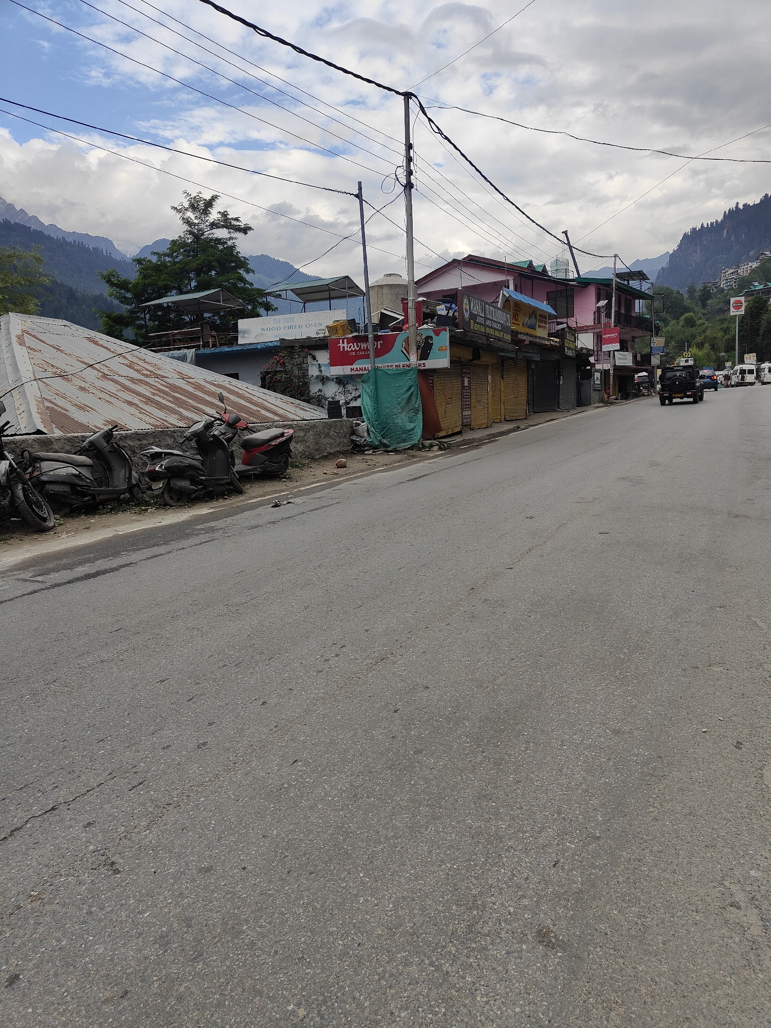 A slightly overcast day in Vashisht, India, shows a view of a street lined with businesses and a backdrop of mountains. Several parked scooters, some appearing worn, are situated along the roadside, near a corrugated metal roof. The businesses display various signs, including "HAVMOR ICE CREAM" and "OVEN IN THE GARDEN WOOD FIRED OVEN." A dark SUV is seen driving away from the camera, followed by other vehicles. The road itself is paved and shows signs of wear. Power lines are visible crisscrossing the sky. The environment suggests a town in a hilly or mountainous region, possibly a tourist destination.