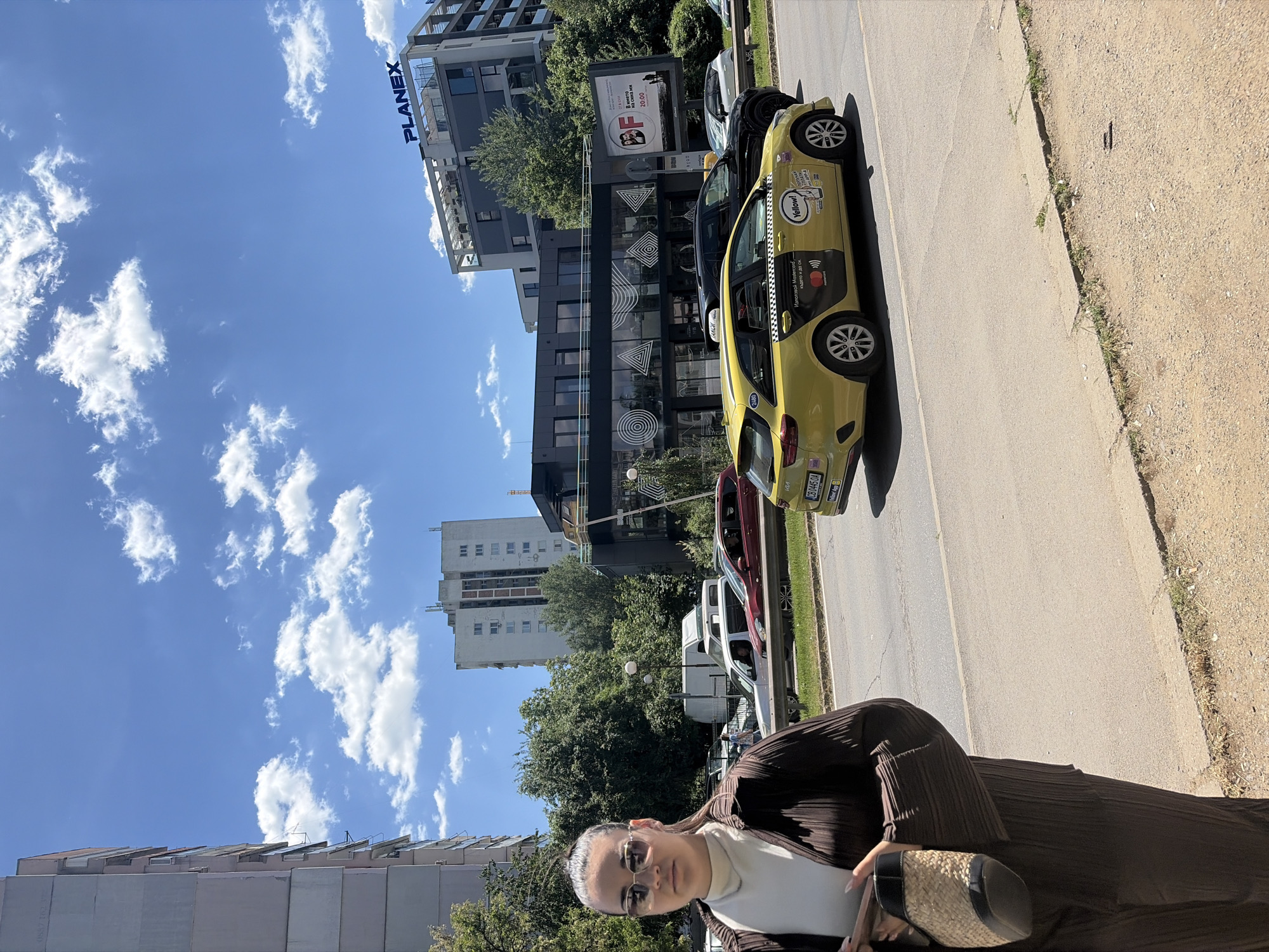 The image depicts a scene in Sofia, Bulgaria. The person in the foreground is wearing sunglasses, a white turtleneck, and a brown pleated outfit while holding a handbag.
A yellow taxi is parked on the side of the road. The taxi has "Yellow!" branding and information on Mastercard payment options in Bulgarian, as well as a license plate number "CB 0445 OA."
In the background are several buildings, including one with the name "PLANEX" displayed prominently. There's also a sign advertising a film titled "В името на сина ми" (In the Name of My Son), with a showing time of 20:00. The sky is blue with scattered clouds.