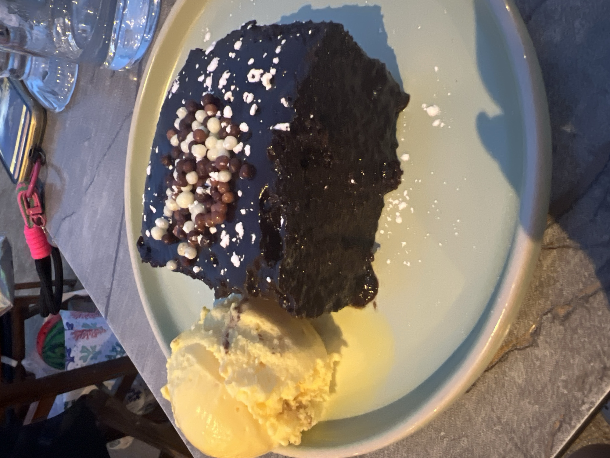 A slice of chocolate cake topped with chocolate and white pearls, alongside a scoop of vanilla ice cream, sits on a pale blue plate. The cake is moist and has a dark chocolate glaze. The plate rests on a grey, textured surface, possibly a table. In the background, a phone with a pink keychain is visible, along with glassware. The image appears to be taken at an outdoor dining setting, possibly in Eretria Municipal Unit, Greece, given the warm lighting and casual atmosphere.
