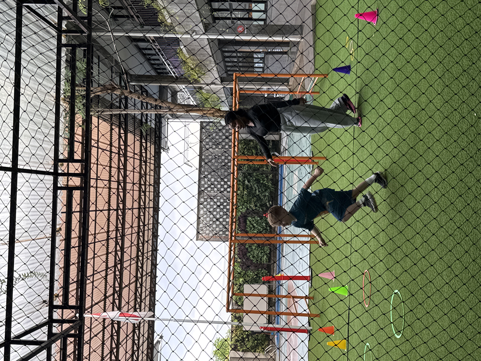 The image depicts an outdoor athletic facility in North Kuta, Indonesia, featuring a green turf field marked with training cones and agility rings. In the foreground, a young Caucasian boy in a dark blue athletic shirt and shorts is running to the right, his body angled towards the camera. Behind him and to his right, a Black woman in a black long-sleeved shirt and grey sweatpants is also running to the right, looking back towards the boy and gesturing with her left hand.
The background includes a multi-story building with balconies and windows, a portion of a swimming pool, and a structure that appears to be a small soccer goal or a piece of playground equipment. A national flag, predominantly white with a red horizontal stripe, is visible on a flagpole. The entire scene is framed by a black netting fence, suggesting the area is enclosed for safety or to prevent balls from leaving the premises. The lighting suggests it is daytime, possibly overcast or late afternoon, with shadows cast on the turf.