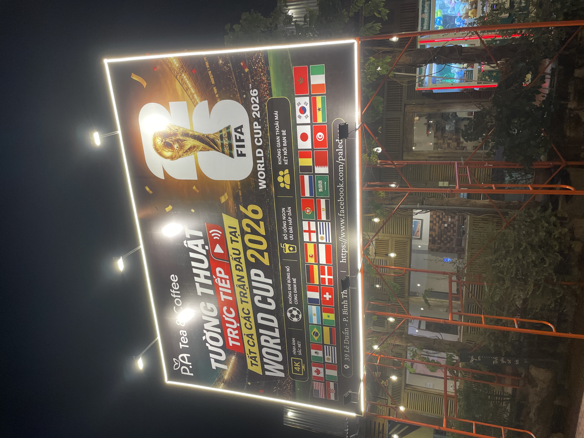 A nighttime view shows a large, illuminated advertisement for the FIFA World Cup 2026 at P.A Tea & Coffee. The sign is in Vietnamese, announcing the live broadcast of all matches at the World Cup. It features the FIFA logo, the World Cup trophy, and a list of participating countries' flags. The sign is located at 39 Lê Duẩn Street in P. Bình Th, Vietnam. The surrounding environment includes outdoor seating, overhead lighting, and some foliage.