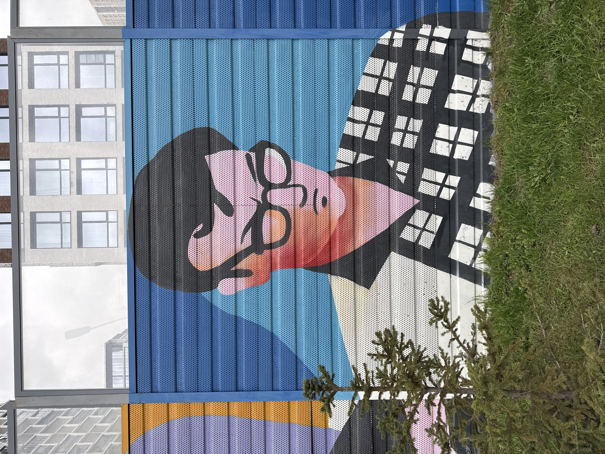 The image is an outdoor shot of an architectural detail. A building facade features a section with a mural depicting a person wearing glasses, rendered in a geometric style. The mural uses shades of blue, pink, black, and white. The person depicted appears to be looking downward.
The mural is installed on a perforated metal panel, revealing the building's support structure beneath. Adjacent to the mural, there are building windows reflecting the sky. There's also a vertical strip of green grass, indicating a planted area.
A small conifer tree branch partially obscures the lower portion of the mural. The building facade is located in Khimki, Russia.