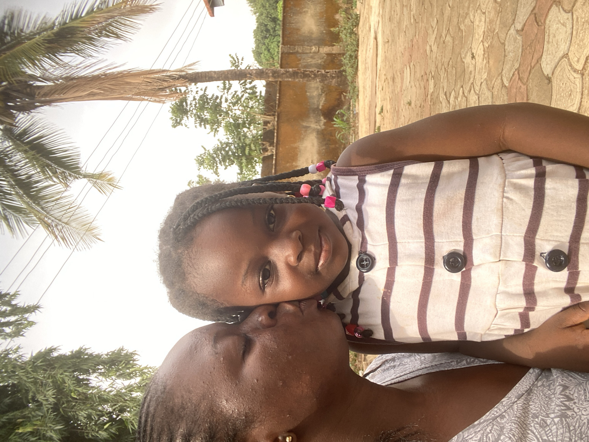 In Maigizo, Nigeria, a woman is kissing a child on the forehead. The child is wearing a striped shirt and has braids with colorful beads. They are outdoors with trees and a wall in the background. The ground appears to be paved.