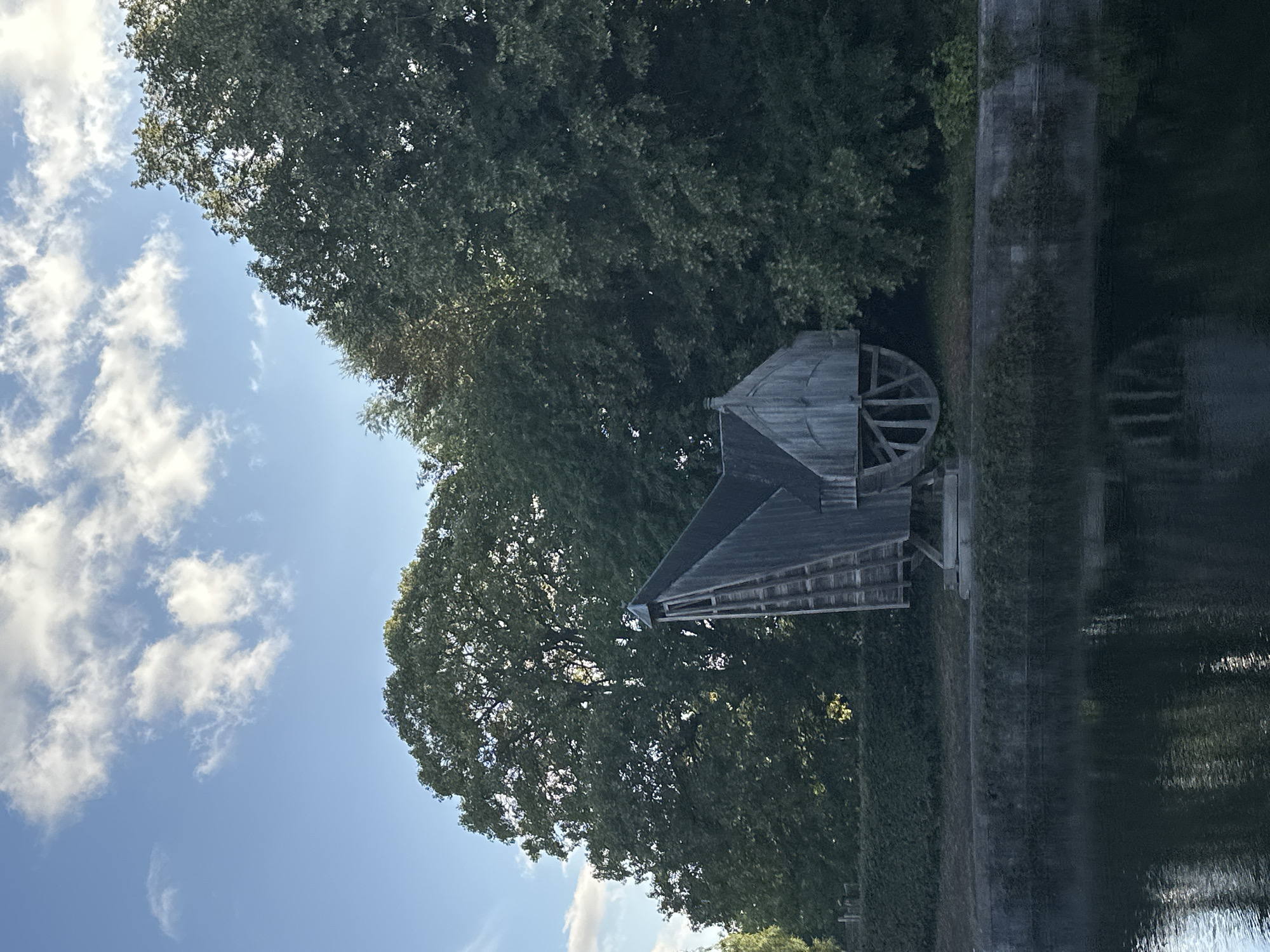 A wooden watermill stands on the bank of a canal in Bruges, Belgium. The watermill is gray and weathered, with a large wooden wheel visible on its side. It is surrounded by lush green trees and a hedge. The sky is blue with scattered white clouds. The water in the canal is dark and reflects the trees and the watermill.