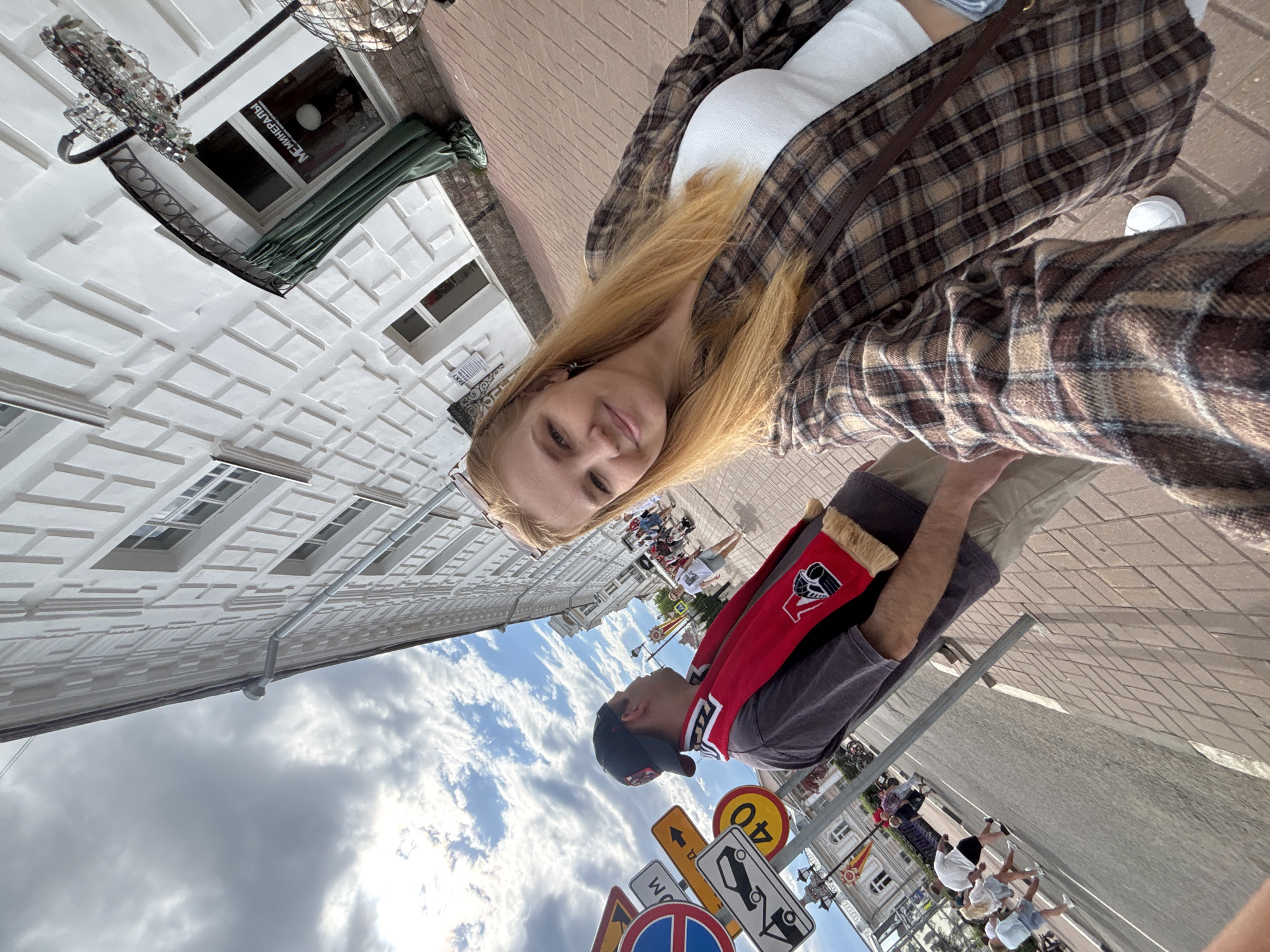 A selfie-style, eye-level shot captures a woman with blonde hair, wearing a white tank top and a brown plaid shirt, posing with a man on a city street. The man is wearing a black baseball cap, a gray shirt, and a red and black scarf. They appear to be standing on a brick-paved street alongside various road signs, against a backdrop of white buildings with ornate architectural details and a cloudy sky. The location is Rusanovo, Russia, evident from the architectural style and street layout. Other pedestrians are visible in the distance, walking on the street.