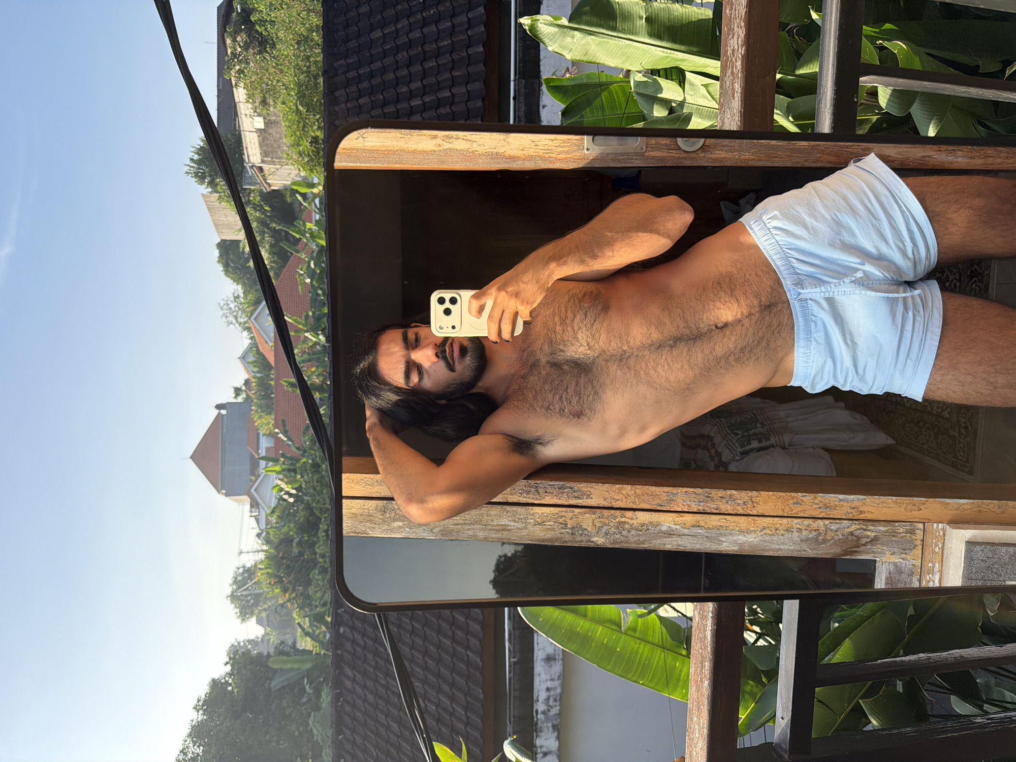 A man with long dark hair and a beard stands on a balcony, taking a selfie in a mirror. He is shirtless and wearing light blue boxer shorts. The man's left arm is raised, with his hand resting on the back of his head. The background shows a residential area with houses and lush green foliage, characteristic of North Kuta, Indonesia. The sun is low in the sky, casting a warm golden light on the scene.