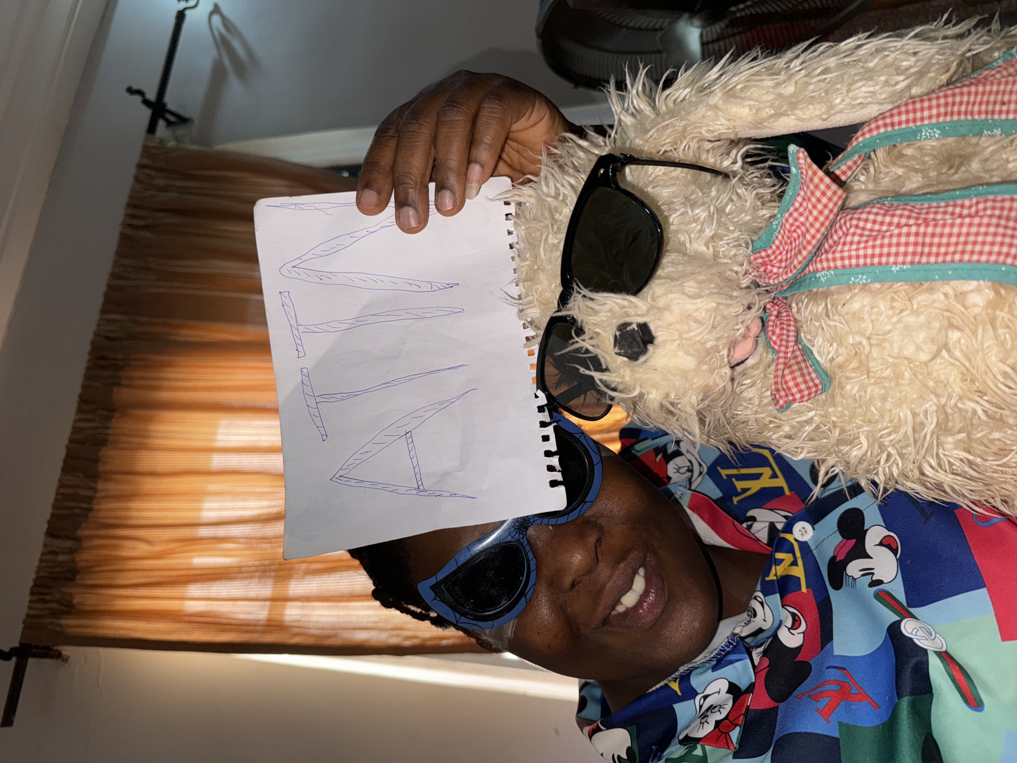 A person is holding a notebook with the handwritten word "NATO" on it. They are wearing sunglasses and a colorful shirt with cartoon characters. A stuffed animal with sunglasses and a red and white checkered bow is next to them. The background includes a window with blinds. The location is Sabon Tasha, Nigeria.