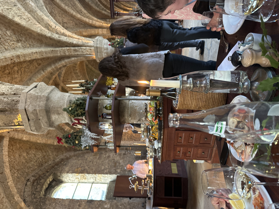 The image depicts an indoor dining or event setting within a large, vaulted stone hall in Cernay-la-Ville, France. The space features exposed light-colored stone walls, a high vaulted ceiling supported by numerous thick stone pillars, and an arched window visible on the far left.
Several pillars are adorned with multi-tiered shelves displaying festive decorations, including small evergreen trees with red bows, silver foliage, and multi-branched candelabras with white candles.
In the midground, a buffet is set up with two dark wooden counters. These counters hold an array of food items in silver serving dishes, chafing dishes, and tiered stands, including various salads and other dishes. A man in a white chef's jacket stands behind the left buffet counter, appearing to attend to the food. Another man, wearing a beige jacket, is partially obscured by a stone pillar near the center buffet. In the midground-right, two women are seen from behind, standing or moving near the buffet area; one wears a light grey textured vest, and the other wears a dark, possibly faux-fur, jacket.
The foreground shows a wooden dining table set with white plates, multiple wine glasses containing white wine, a clear glass bottle of Badoit mineral water, and a silver candlestick holding a lit white candle. A plate on the table contains seafood, specifically prawns and crab claws, accompanied by lemon wedges. A person's hand is visible on the left, near a plate, and a male diner is partially visible on the right, holding a glass. The scene is illuminated by ambient light and the flickering candle flame.