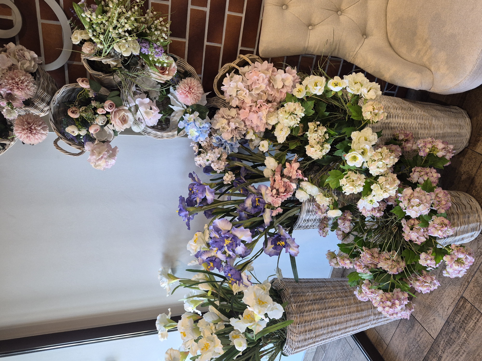 A collection of wicker baskets filled with floral arrangements is displayed against a brick wall and part of an upholstered chair. The flowers vary in color, including pink, white, purple, and blue. The baskets are arranged on a wooden floor and a white surface, possibly a table. There is no indication of the location of the scene in Votkinsk, Russia. No people are visible.