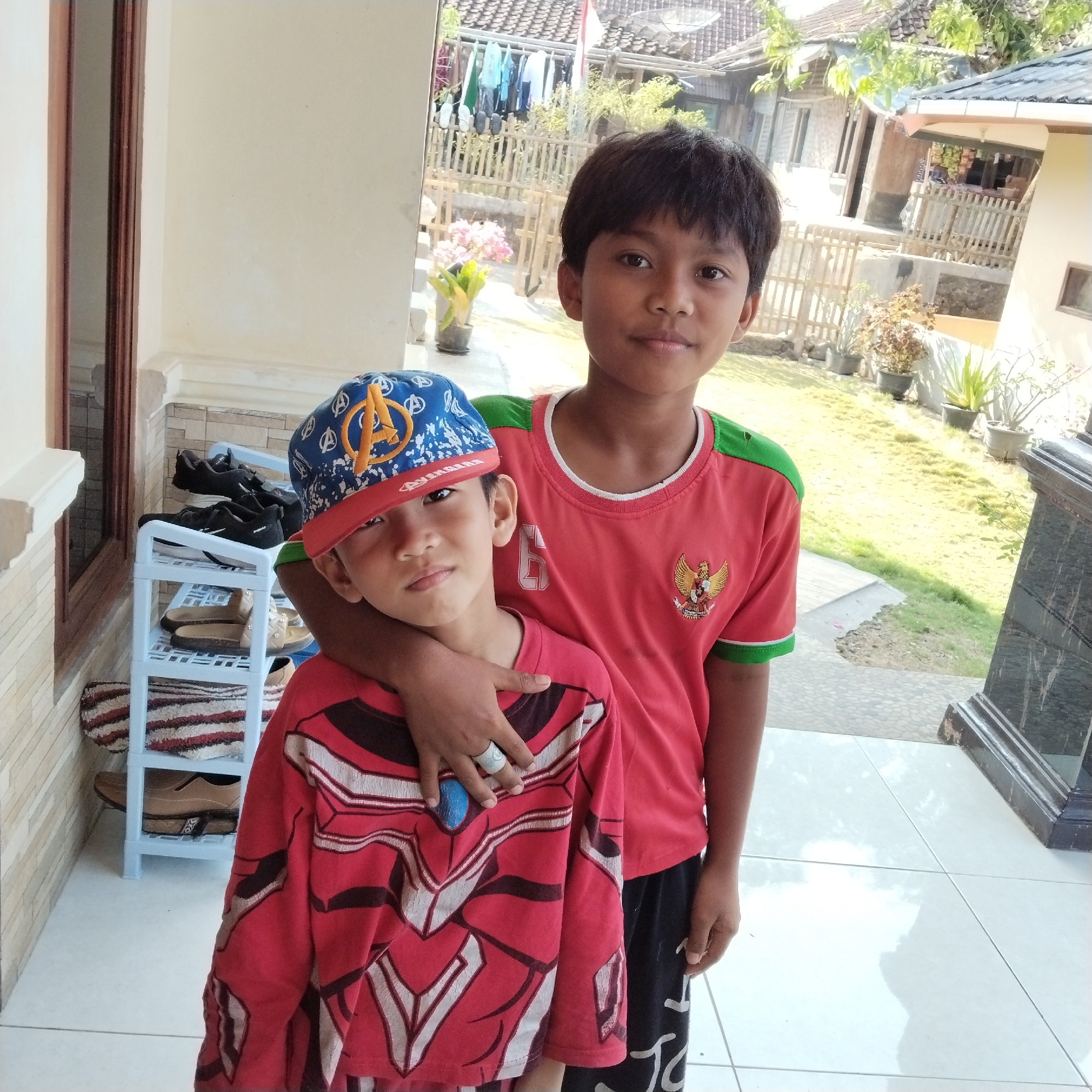 Two young boys stand on a tiled porch with a background of a residential area. The boy on the left wears a red shirt with a superhero design and a blue baseball cap with an Avengers logo. The boy on the right wears a red t-shirt with green trim and a Garuda emblem. The older boy has his arm around the younger boy's shoulders. In the background, a shoe rack with several pairs of shoes is visible. The environment appears to be outdoors, with a lawn, plants, and simple houses suggesting a rural or suburban setting. The sunlight indicates it is daytime.