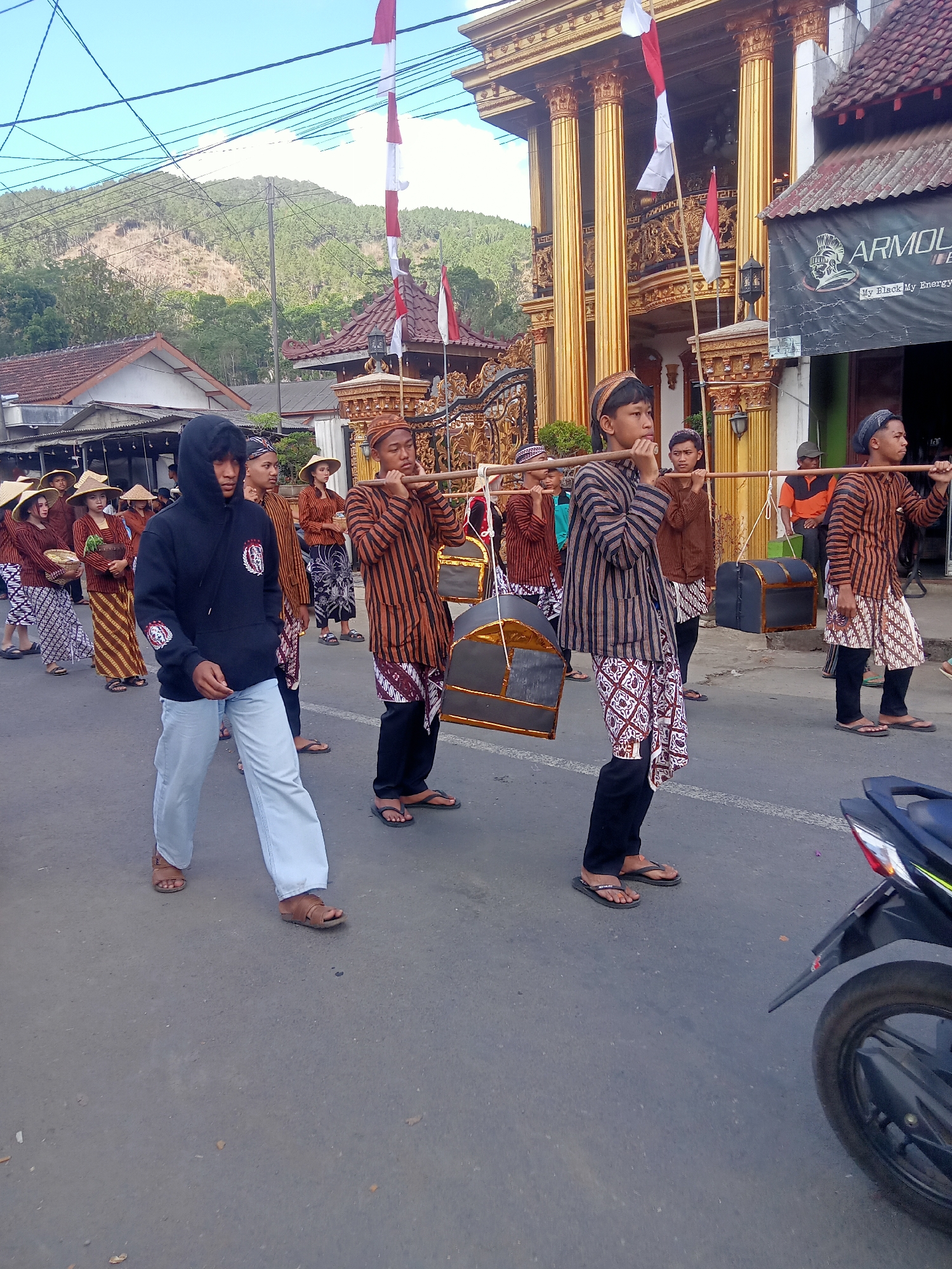 In the city of Pule, Indonesia, a parade is underway on a paved street. The parade features a group of people, primarily young men and women, dressed in traditional Indonesian attire. Many of the participants wear striped shirts, patterned sarongs, and hats. Several young men are carrying large, dark-colored chests or containers suspended from wooden poles balanced across their shoulders. A young man in the foreground is wearing a black hoodie and light-colored pants. In the background, a large, ornate building with golden columns and red-tiled roofs is visible. The sky is blue with some clouds, and a line of trees and hills can be seen in the distance. A motorcycle is parked on the right side of the street, partially visible in the frame. The scene appears to be a cultural or religious procession.