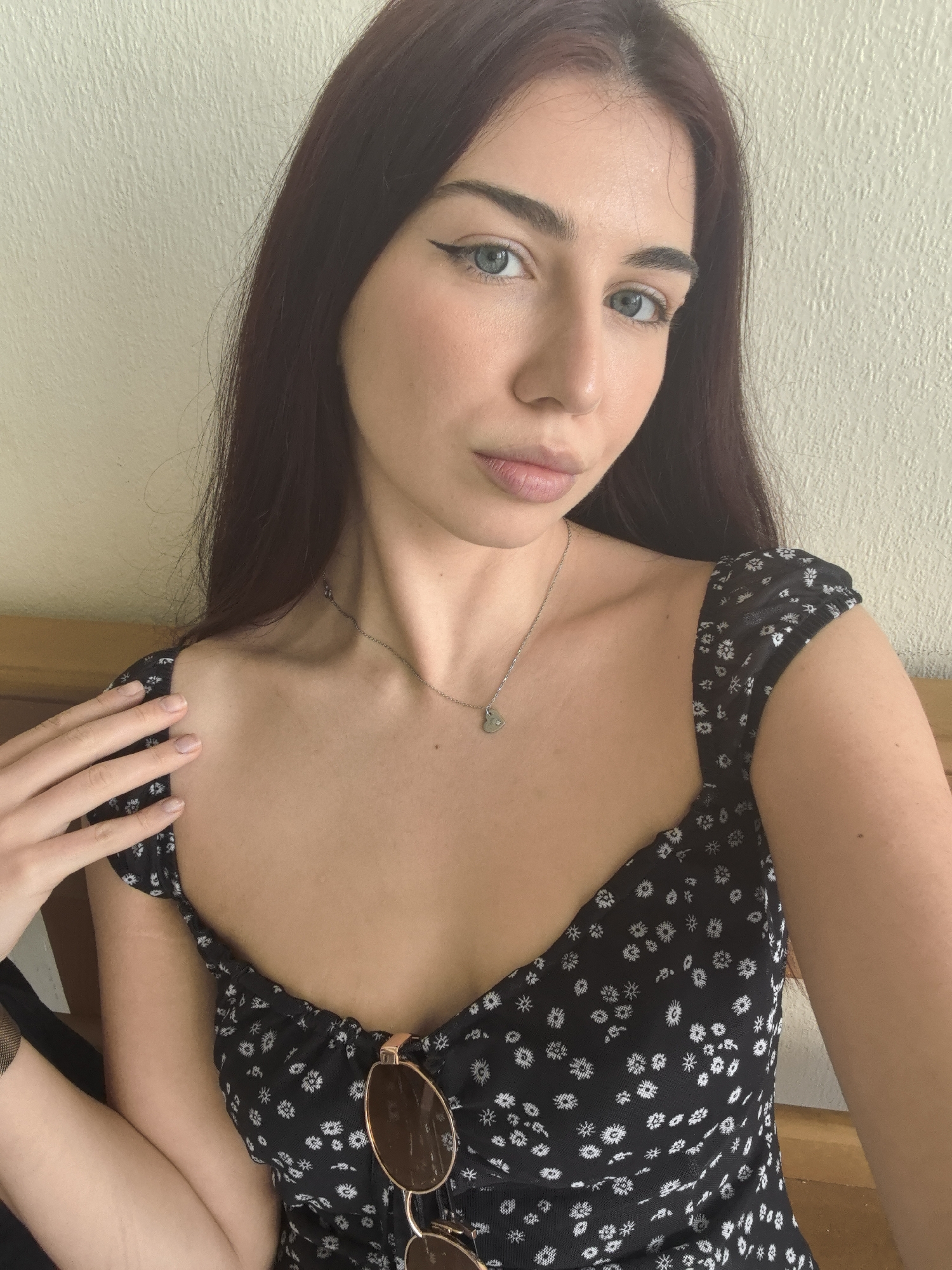 A head-and-shoulders selfie captures a young woman with long, dark reddish-brown hair and striking light blue or grey eyes. She is looking directly at the camera with a neutral expression. Her left eye is enhanced with a distinct winged black eyeliner, while her right eye has a more natural look. Her eyebrows are neatly shaped and full. She wears a black top with small white floral patterns, featuring a sweetheart neckline and ruched cap sleeves.
Around her neck is a delicate silver chain necklace with a small, possibly heart-shaped or irregular charm. A pair of round, brown-lensed sunglasses with thin gold frames is tucked into the front of her top. Her left hand, with natural-looking nails, rests gently on her right shoulder.
The background consists of a light-colored, textured wall. A portion of a light wooden structure, possibly furniture, is visible to the left and behind her. The scene is illuminated by natural daylight, indicating it is daytime. The overall setting appears to be an indoor, casual environment in Stara Zagora, Bulgaria. No text is visible within the image.