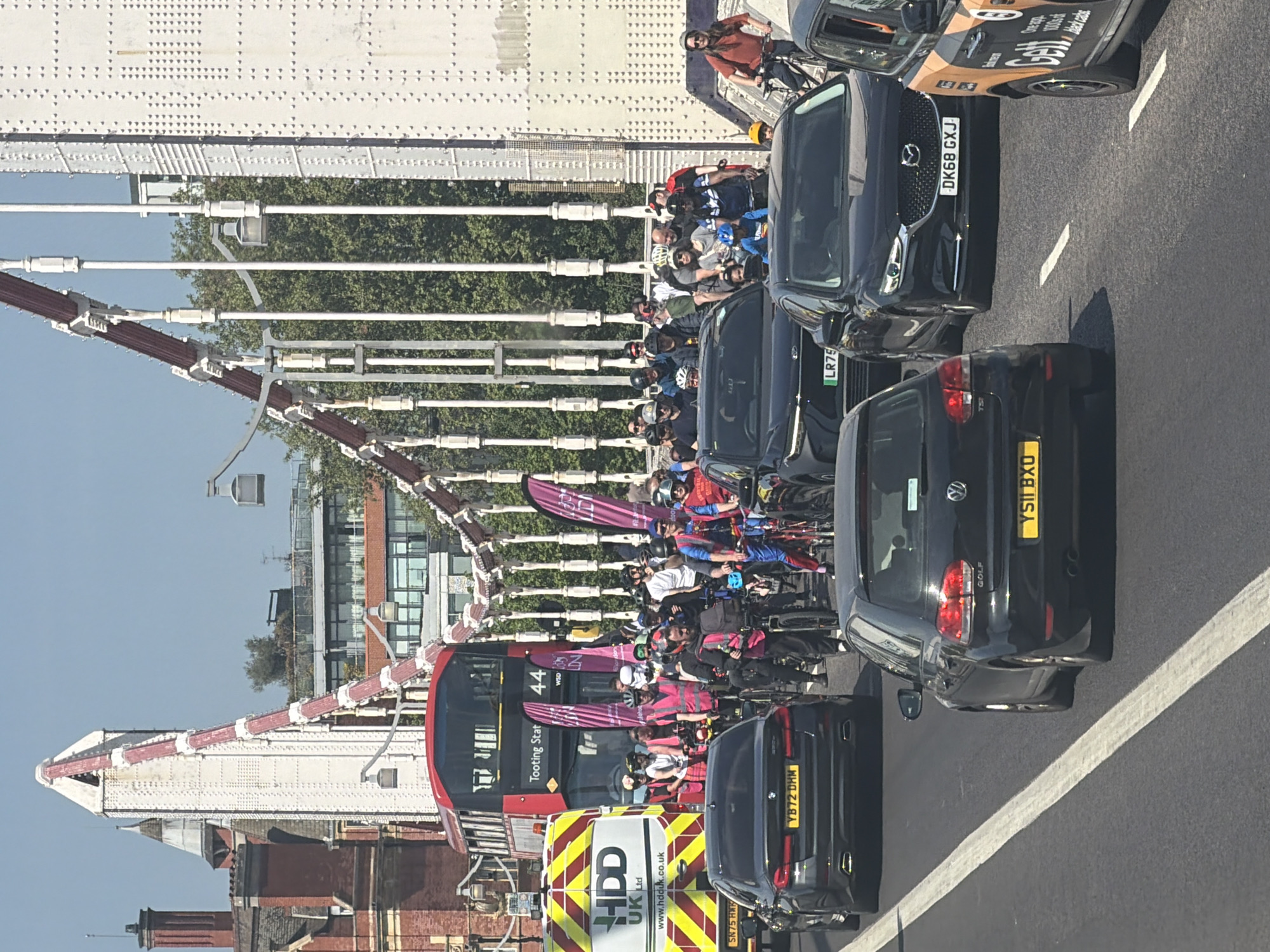 The image captures a view of a street in London, United Kingdom, heavily congested with traffic and pedestrians. The scene appears to be set on a bridge, evident from the large white steel structure overhead. A significant number of pedestrians, many on bicycles and potentially part of an organized event or ride, are situated on the left side of the road, with vehicles, including cars, a black cab, a double-decker bus (route 44 to Tooting Station), and a van with the branding "HDD UK Ltd," occupying the right side. Various car license plates are visible, including DK68 GXJ, YS11 BXO, LR75, SN75 HWK, and YB72 DHM. The environment includes buildings and trees in the background, suggesting an urban setting. The sky is light blue, indicating a sunny day.