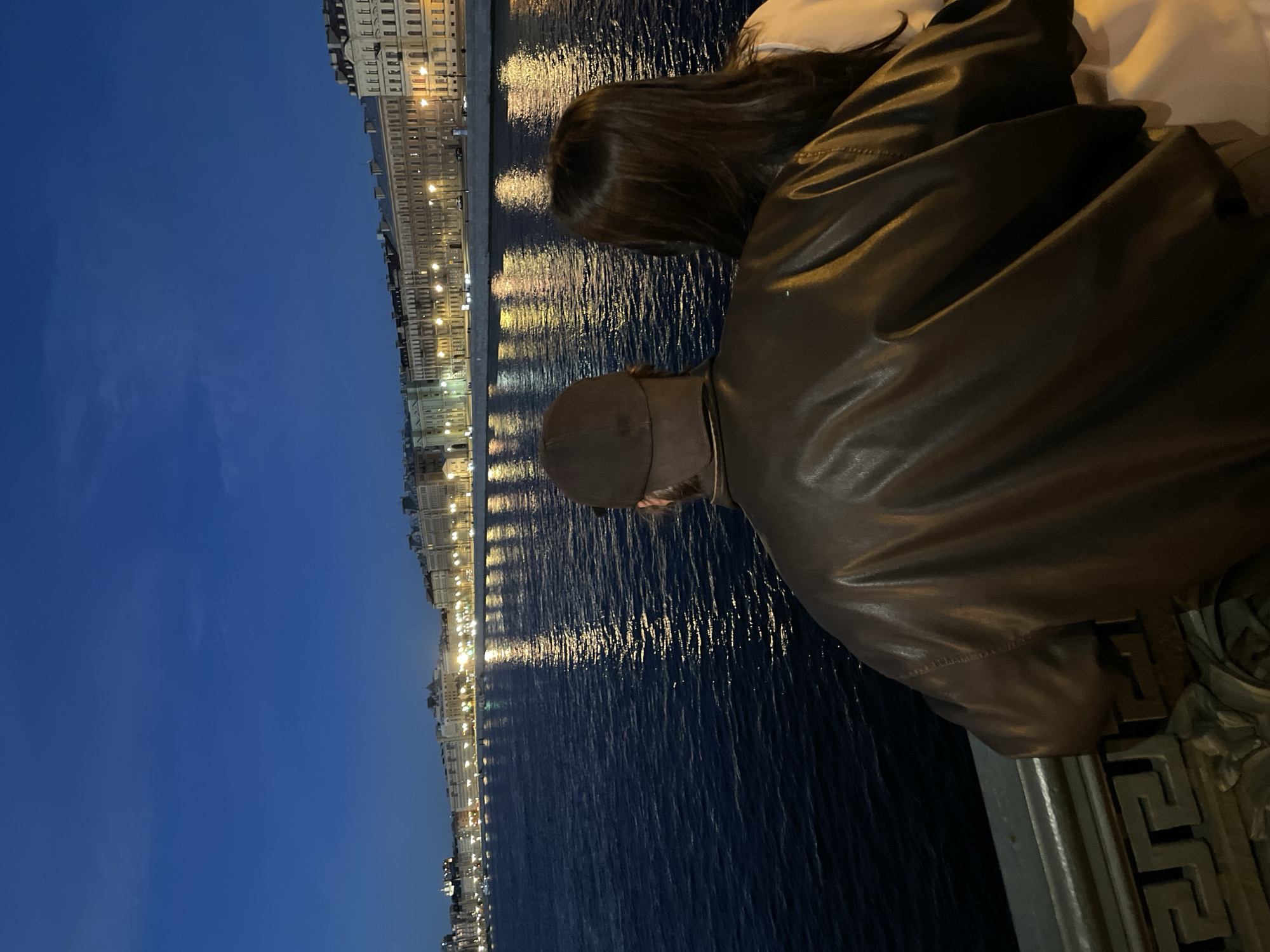 This is a nighttime, medium shot of two people from behind, looking out at a river and city skyline. The person in the foreground on the left is wearing a brown leather jacket and a brown baseball cap. The person next to them on the right has long, dark hair and is wearing a light-colored top. They are standing on a bridge or promenade, with an ornate metal railing in the lower part of the frame. The river is dark and reflects the lights of the buildings on the opposite bank. The buildings are lit up with warm yellow lights, creating a soft glow. The sky is a deep blue, indicating it is late evening or early morning. The overall mood is serene and romantic, with a sense of quiet contemplation.