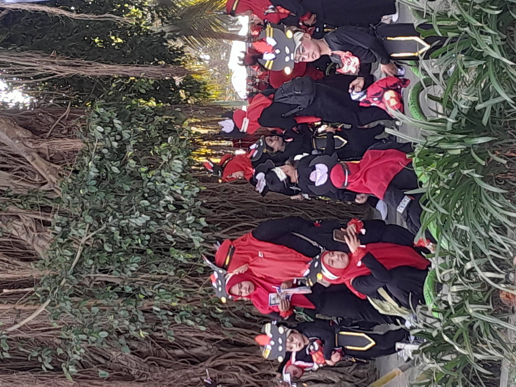 In Genteng Kulon, Indonesia, a group of people, including several children, are gathered outdoors in front of a large tree with hanging aerial roots. Many of the individuals are wearing matching black and red costumes with elaborate headpieces that resemble animal or mythical creatures, featuring large, stylized eyes and horns. The costumes for the children include black tops and pants with decorative yellow trim, and some wear masks around their necks. The adults are also dressed in red headscarves and black attire, with some holding items like brochures or phones. The lighting suggests it is daytime, and the presence of lush green plants in the foreground indicates a park or garden setting. Some individuals are interacting, with one woman in the foreground looking at her phone while seated. The overall scene appears to be a gathering for a performance or cultural event.